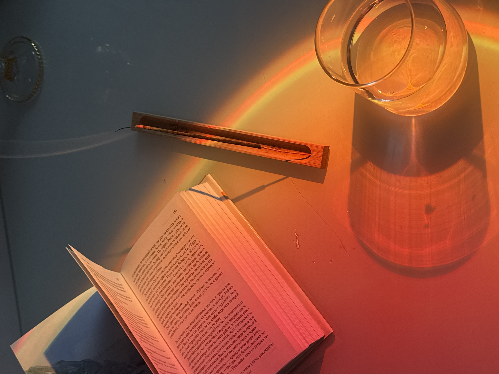 A high-angle shot presents a scene of leisure and study bathed in warm, colorful light. The image features an open book, positioned at the lower left. The pages, displaying text in a non-English language, suggest an ongoing reading session. Adjacent to the book, a wooden incense holder lies horizontally, with an incense stick placed inside, indicating a possible scent. A clear glass water pitcher is placed on the upper right. The entire setting is illuminated by a rainbow of color, cast across the objects and the surface. The scene has a calm and relaxing feel.