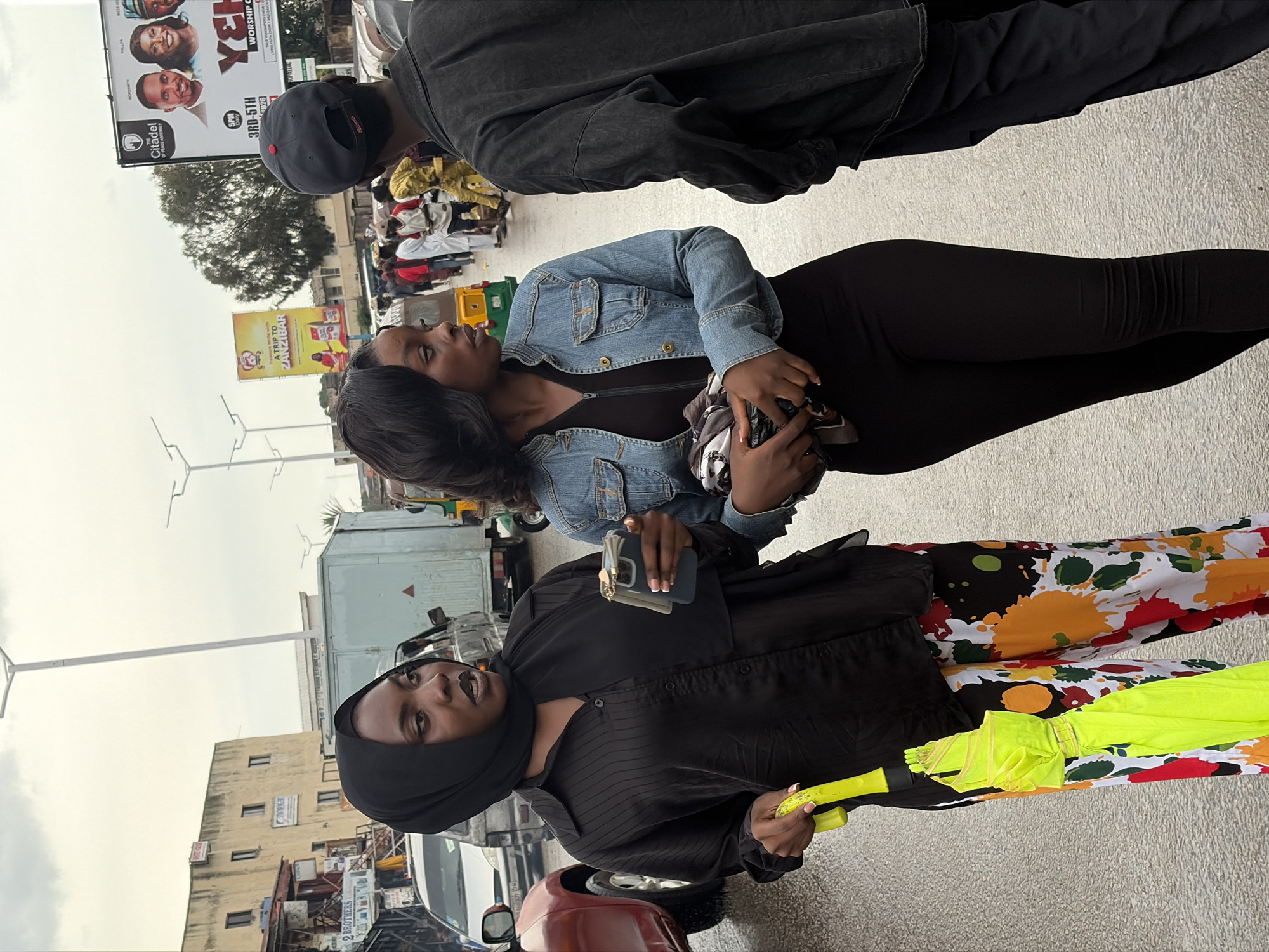 The image shows a group of people standing on a street. In the foreground, two women are visible. The woman on the left is wearing a black headscarf and a black button-up shirt, holding a yellow umbrella. The woman on the right is wearing a denim jacket over a black dress and is holding a phone. Behind them, another person in a dark jacket is partially visible.
The setting appears to be an urban environment, likely in Jos, Nigeria, with buildings and streetlights in the background. A billboard advertising "Citadel" and "WORSHIP C" is prominent, along with another billboard for a "TRIP TO ZANZIBAR." The weather seems overcast. The overall mood is casual, with the subjects looking relaxed. The time of day is not explicitly clear but appears to be daytime.
