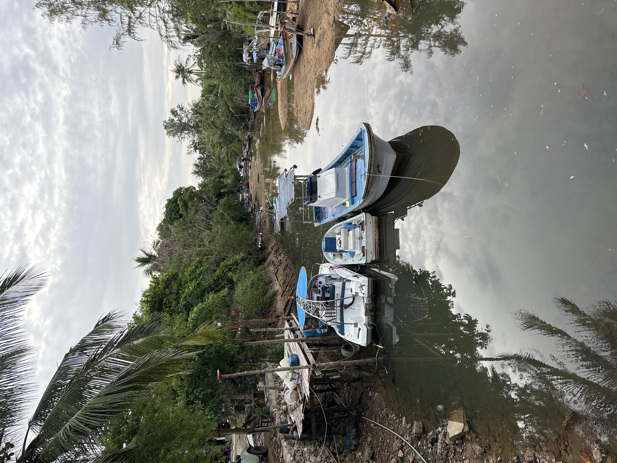 An outdoor, slightly elevated view captures a serene waterway lined with lush tropical vegetation under an overcast sky. The image is set in what appears to be a small inlet or river, characteristic of a coastal or island environment like Ko Pha Ngan, Thailand.
In the foreground and middle ground, several small boats are visible. Some, including a prominent light blue and white boat with a cabin and what appears to be a solar panel setup, are moored in the calm, reflective water. Another white boat with a blue-and-white interior and a prominent blue dish antenna is also moored near a rickety wooden pier or platform. This platform, made of planks and supported by poles, has various items on it, including containers and what looks like discarded materials, suggesting a working or residential dockside area. The water itself reflects the cloudy sky and the surrounding trees, with some leaves floating on its surface.
Along the sandy-muddy bank to the left, several other small, colorful fishing boats are either pulled ashore or partially submerged at the water's edge. One red, white, and blue boat prominently displays the number "8" and the faded text "K. Champ" or "King Champ" on its bow. The banks are densely covered with a variety of green trees, including tall palm trees, their fronds visible against the sky. The sky is predominantly cloudy with varying shades of white and gray, indicating an overcast day. No people are visible, and the scene conveys a quiet, tranquil atmosphere, likely representing a local fishing or boat docking area.
