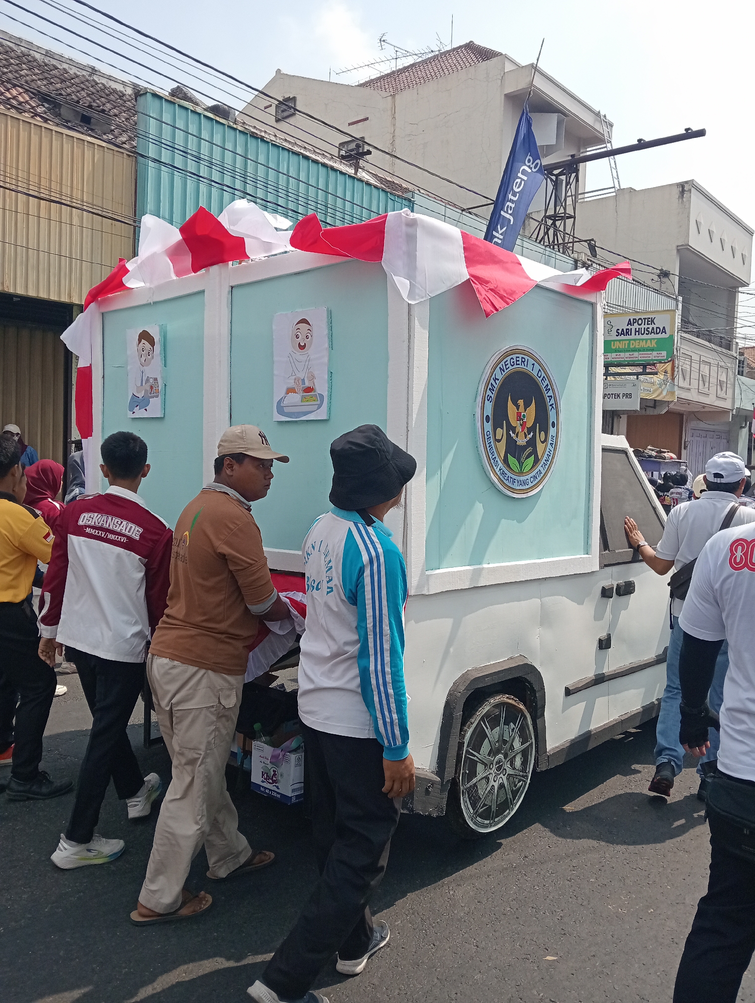 A group of people are walking on an asphalt street in what appears to be a procession or parade. In the foreground, several individuals are visible from the waist down, some wearing casual clothing like t-shirts, pants, and sneakers, while others are in more traditional attire. A significant element of the scene is a large, white box-like structure mounted on a vehicle, decorated with red and white streamers and two framed images. A circular emblem with the text "SMK NEGERI 1 DEMAK" and "GENERASI KREATIF YANG CINTA TANAH AIR" is prominently displayed on the side of this structure. In the background, buildings with various architectural styles line the street, and electrical wires are strung overhead. A blue flag with white text "k Jateng" is visible on a pole. The overall atmosphere suggests a public event or celebration taking place in a moderately populated urban or suburban area.