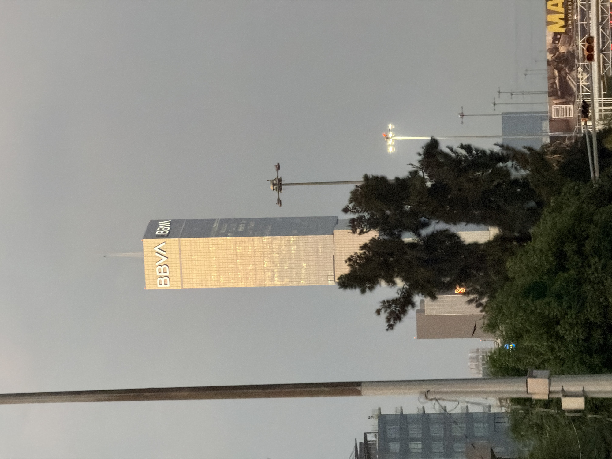 The image shows a cityscape in San Bernardino Tlaxcalancingo, Mexico under an overcast sky. The BBVA building is a prominent feature, towering over other structures. Utility poles are visible, and trees are present in the foreground, partially obscuring some buildings. A billboard can be seen on the right side of the frame.