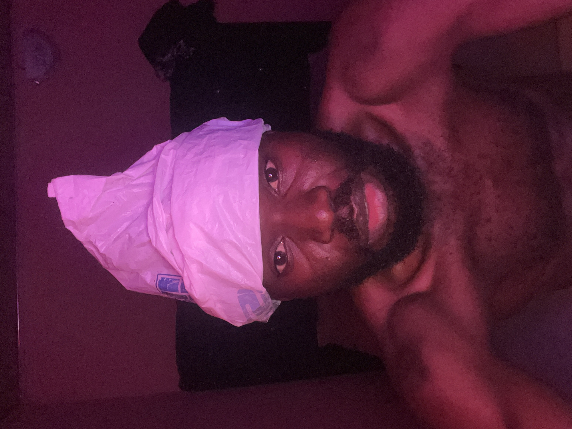 A close-up, low-angle shot depicts a man with a dark complexion, beard, and exposed chest. He is positioned with a plastic bag covering his head. The background is dimly lit with a red hue and includes a black T-shirt. The location of the image is not discernible as Gura Top, Nigeria.