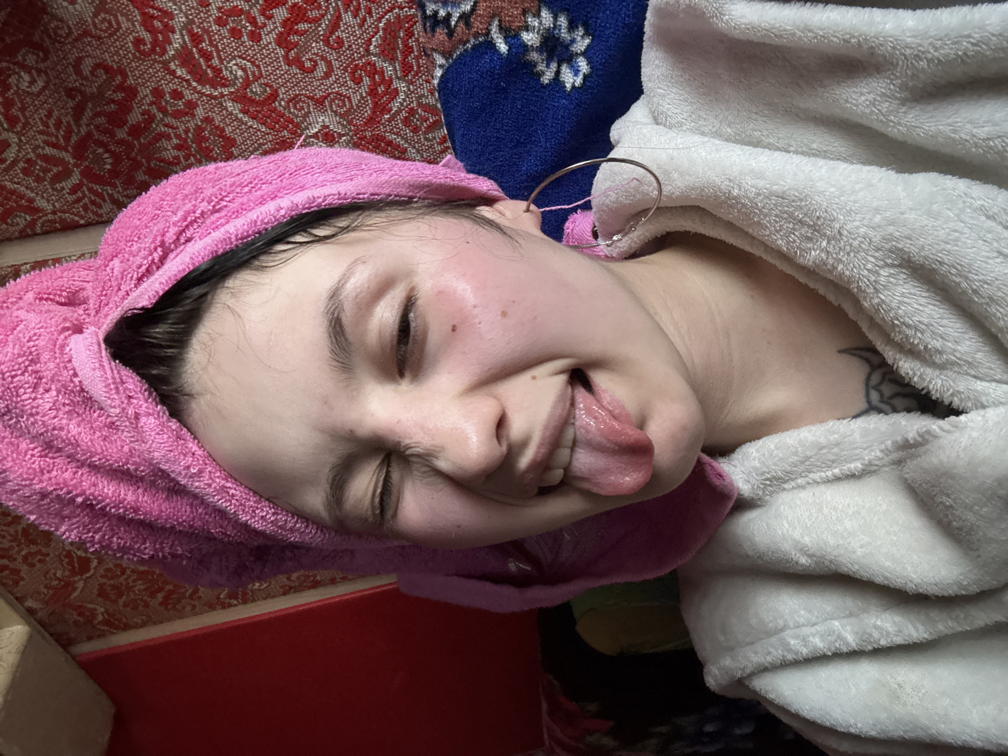 A young individual is lying down with a pink towel wrapped around their head and a white robe draped over them. They are sticking out their tongue and squinting their eyes. The background includes a patterned red and brown fabric and a blue floral pattern. A red surface is visible in the lower left corner. The location is unknown and cannot be determined from the image.