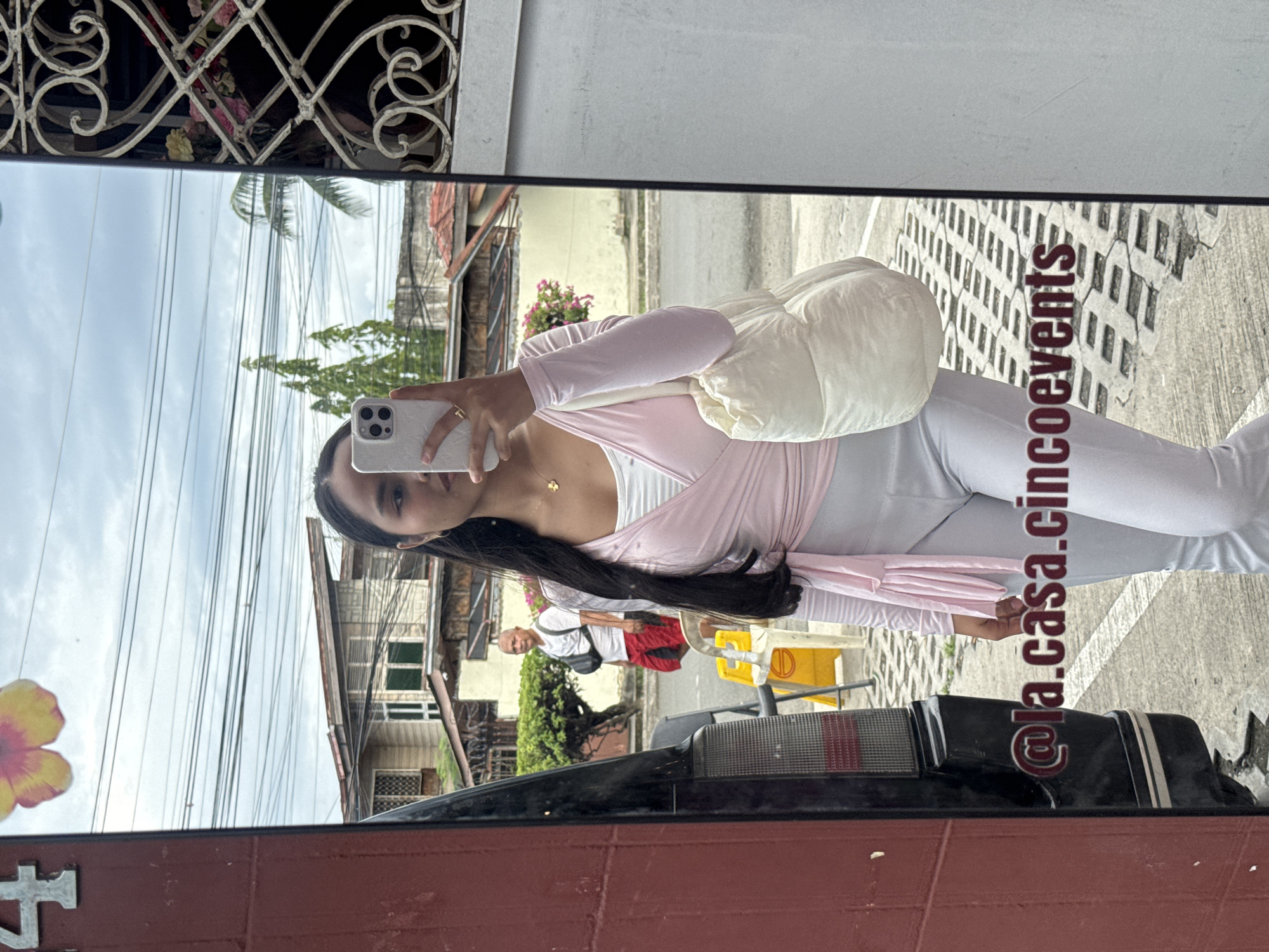 A young woman is reflected in a car side mirror, taking a selfie with a white smartphone. She is wearing a white top with a pink wrap or cardigan, and light-colored pants. She has long, dark hair and is wearing a gold necklace. The reflection shows an outdoor scene, possibly a street in Davao City, Philippines. The background includes houses, utility poles, vegetation, and people walking in the distance. A yellow and white object is partially visible behind the car. The mirror is attached to a dark-colored vehicle with visible taillights. The foreground includes a red brick or painted surface. Text, "@la.casa.cincoevents", is superimposed vertically in the image. A wrought iron pattern is visible in the top left corner. A flower is shown next to the edge of the mirror.