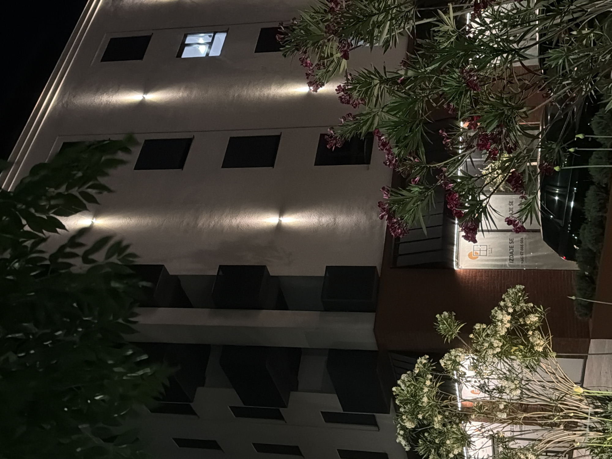 Here is a detailed description of the image:
**Setting:** The photograph appears to be taken at night in Podgorica, Montenegro, based on the advertising signage on the building.
**Building:** It features a modern, multi-story building with a light-colored facade and dark windows. The facade has recessed window areas creating architectural details. There are lights mounted on the exterior of the building, illuminating the facade in sections.
**Vegetation:** Several trees and bushes are in the foreground, partially obscuring the view of the building. The vegetation includes what appears to be an oleander with pink blossoms and another plant with white flowers.
**Additional Details:** A section of the building's lower level has a different architectural style, possibly brick or textured paneling, and features signage with a telephone number. A vehicle is partially visible, seemingly parked alongside the building.