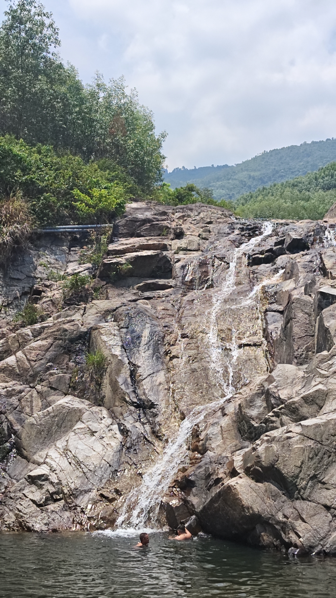 A daytime exterior shot captures a waterfall cascading down a rocky slope into a body of water. Two individuals are partially submerged in the water at the base of the waterfall. The waterfall's flow is concentrated, creating a white stream against the dark rocks.
The rocky slope is composed of large, angular stones. Vegetation, including trees and green foliage, is visible at the top of the rocks and on the left side of the frame. The background features mountains covered with trees under a cloudy sky.
The location is in the city of Đà Nẵng, Vietnam.