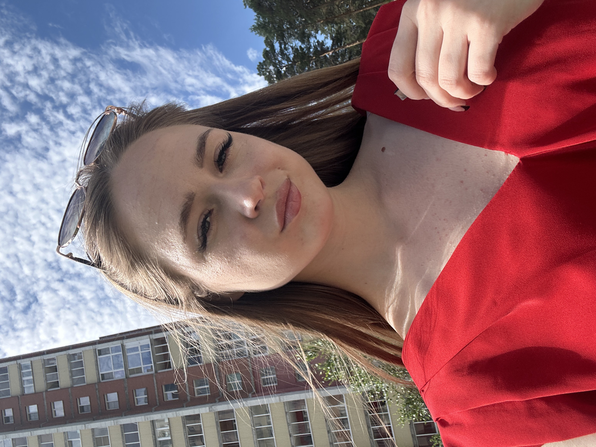 A young woman with long brown hair, wearing a red dress and sunglasses, poses for a selfie outdoors. She is standing in front of a tall apartment building with a blue sky and clouds in the background. A portion of a forest is visible to the right of the building. The woman's left hand is visible near her chest, and she appears to be holding something.