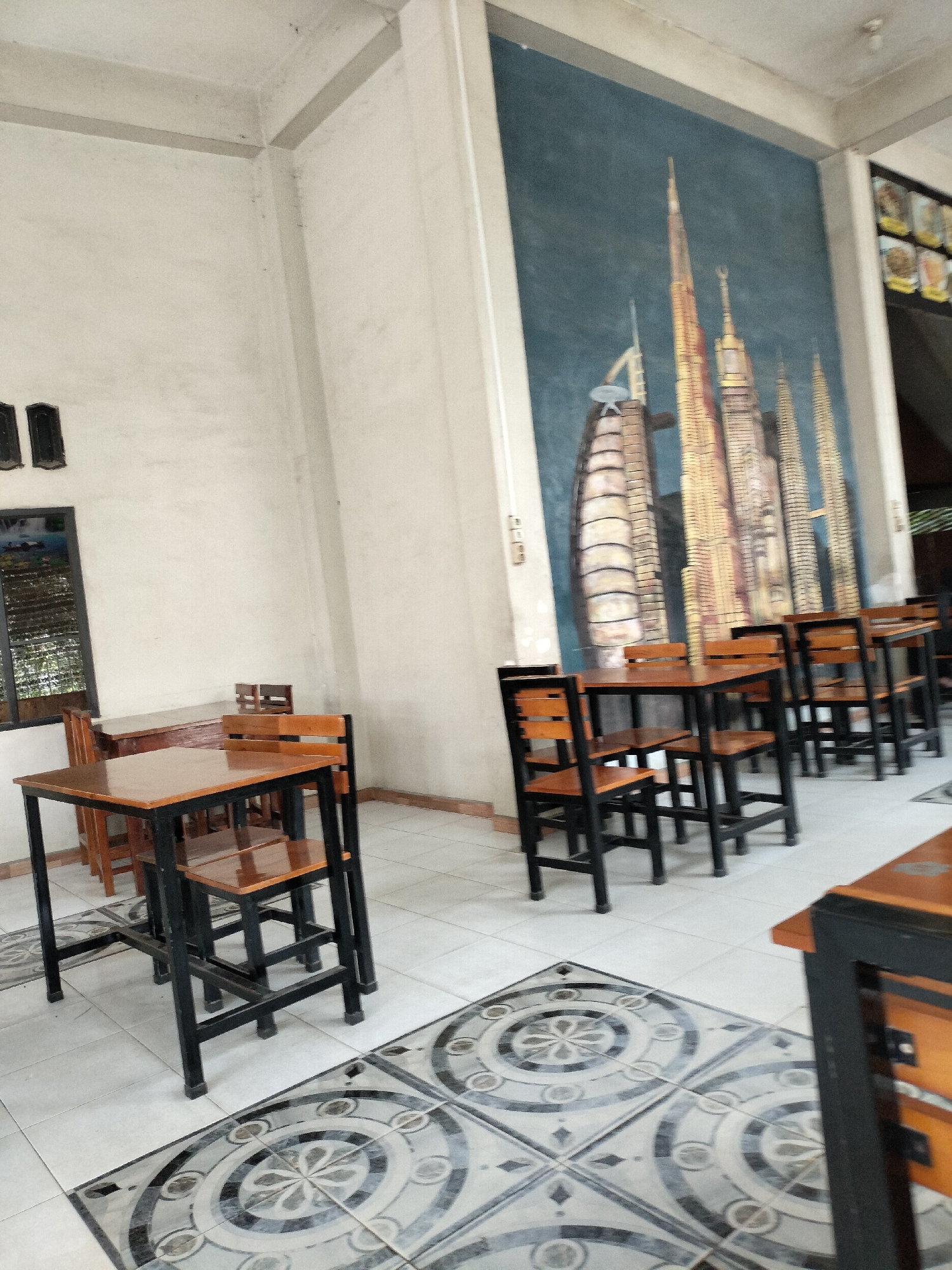The image depicts the interior of a restaurant in Keudealue Ie Puteh, Indonesia. The dining area is furnished with multiple sets of wooden tables and chairs, all featuring black metal frames. The walls are painted a light color, and one wall features a large mural of skyscrapers. The floor is tiled with a decorative pattern. No people are visible in the image. The scene appears to be well-lit, likely due to natural light from unseen windows.