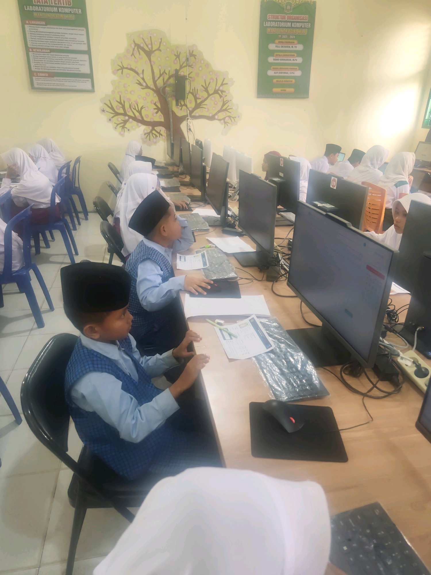 The image shows a computer lab with several children wearing school uniforms, including white shirts, blue vests, and peci hats, seated at desks with computers. The lab appears to be located in Pematang Reba, Indonesia, as indicated by posters on the wall with Indonesian text, specifically mentioning "Laboratorium Komputer MAN Indragirihulu" which translates to "MAN Indragirihulu Computer Laboratory." One poster details "Tata Tertib Laboratorium Komputer" (Computer Laboratory Regulations), and another displays the "Struktur Organisasi Laboratorium Komputer" (Organizational Structure of the Computer Laboratory). The environment is a classroom setting with beige tiled floors and walls adorned with a decorative tree mural. The children are engaged with the computers, some looking at their screens, others with their hands on the keyboards or mice. The overall scene depicts an educational activity in a computer lab.