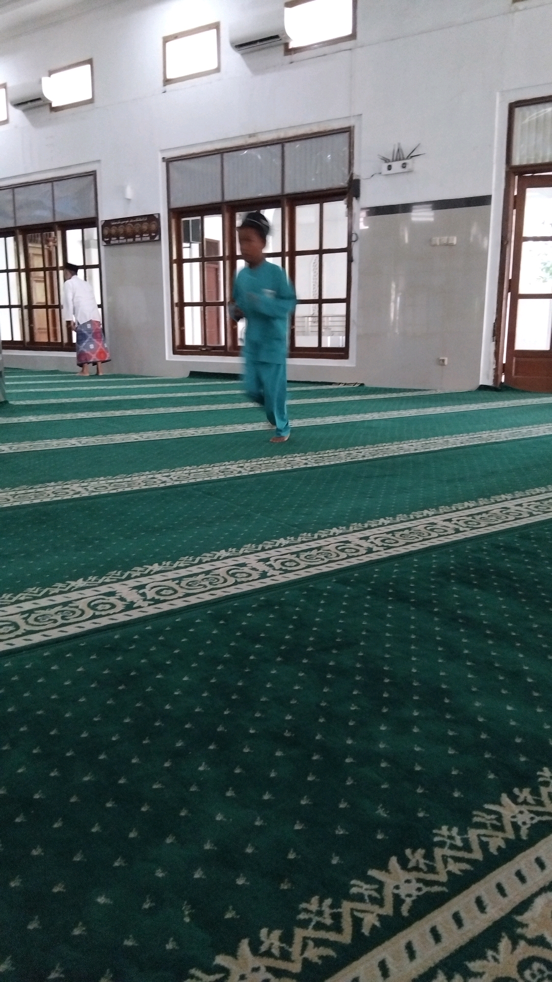 In the city of Batang, Indonesia, a boy in a teal jumpsuit and cap is captured in motion, running across the green carpeted floor of what appears to be a mosque. The carpet is marked with white striped patterns. To the left, a person in a white shirt and patterned sarong stands near a window. The room features white walls, wooden-framed windows, and air conditioning units mounted high on the wall. The lighting in the mosque is bright, illuminating the space.