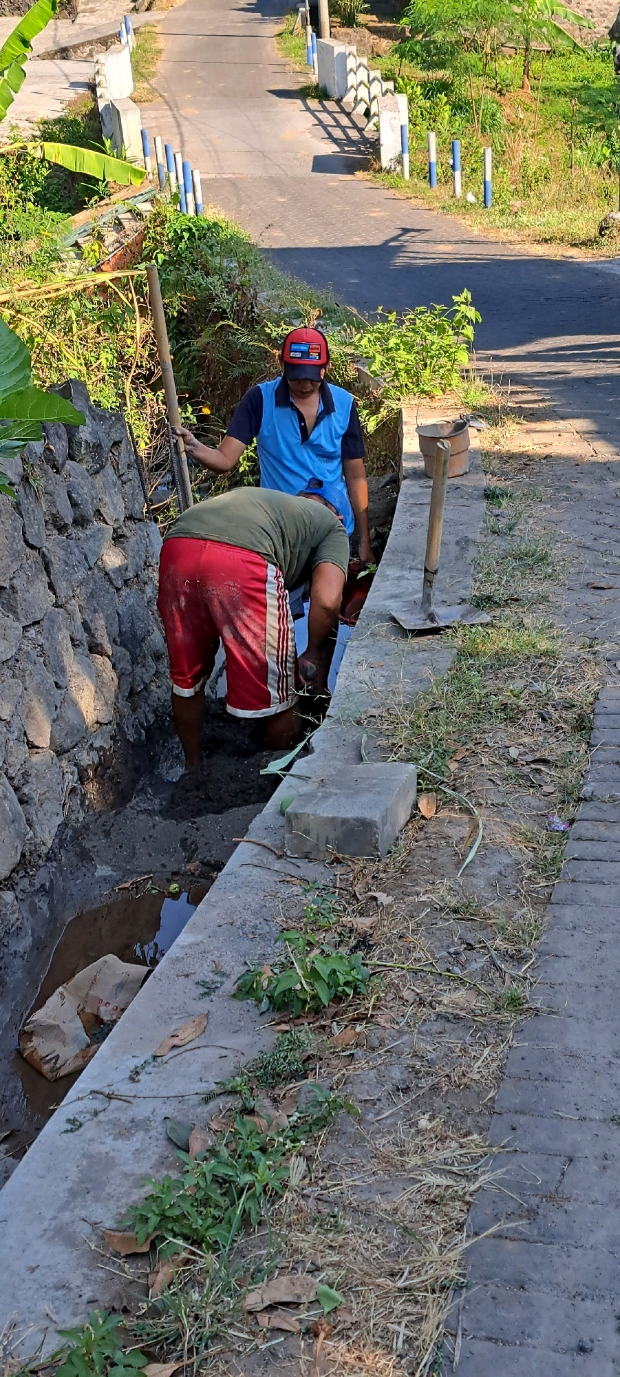 Two men are working in a drainage ditch next to a road in Pandaan, Indonesia. One man, wearing red shorts and a green shirt, is bending over in the ditch, digging with his hands or a tool. The other man, wearing a blue shirt and a red cap, stands behind him, holding a long pole or shovel. The ditch is filled with muddy water and debris. The road is paved and lined with concrete barriers and vegetation. The scene appears to be a residential area or a rural road.