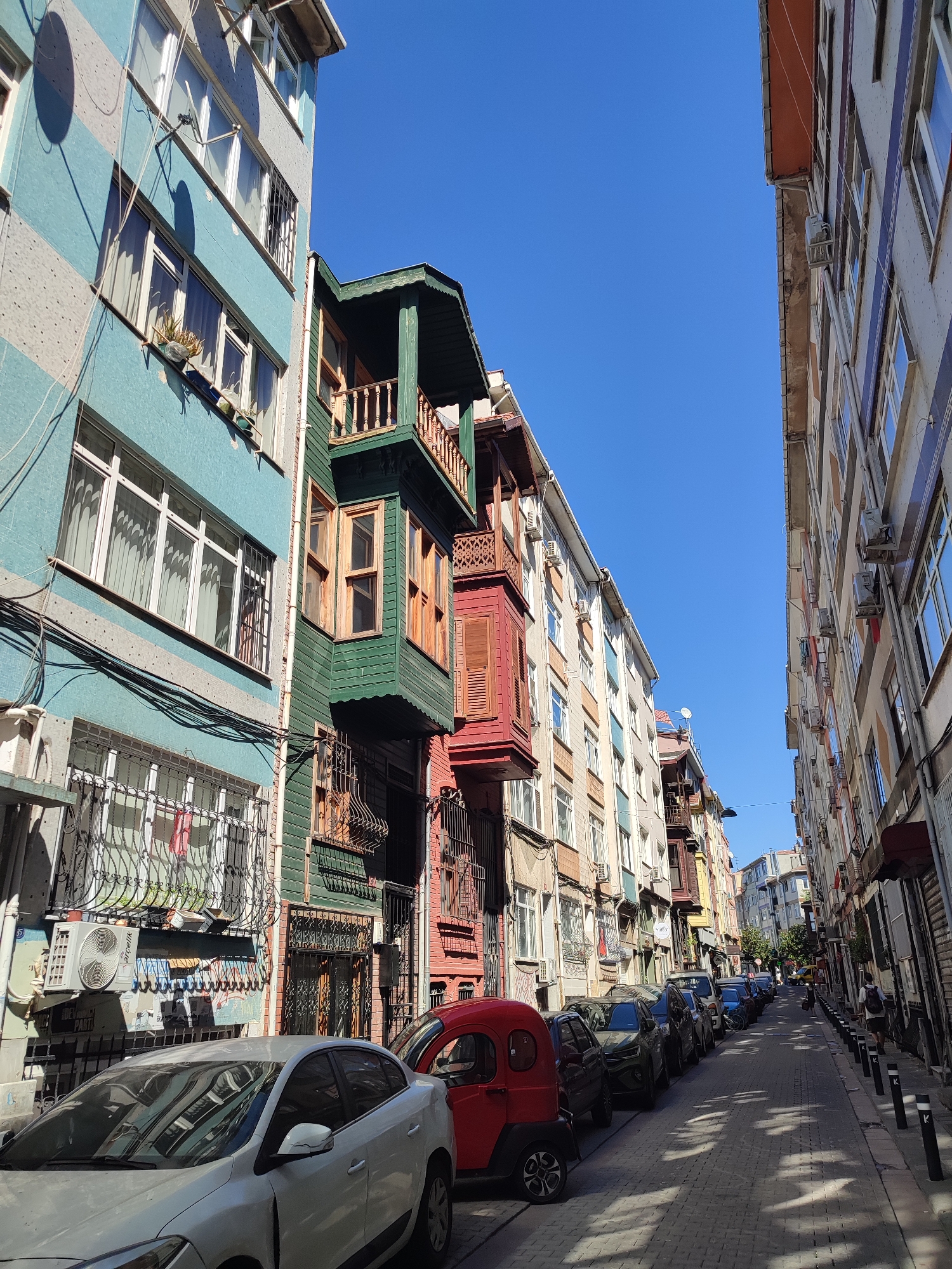 The image depicts a narrow, cobblestone street in Kadıköy, Turkey, lined with multi-story buildings. The architecture is varied, with some buildings painted in bright colors like turquoise and green, featuring traditional bay windows and wooden balconies. Others are more muted in tone, with striped facades and visible air conditioning units. Cars are parked along both sides of the street, with a white sedan and a small red vehicle prominent in the foreground. A lone pedestrian, a person with a backpack, walks down the right side of the street. The sky is clear and blue, indicating a sunny day. Shadows from the buildings and trees are cast across the street, creating a dappled effect on the cobblestones.