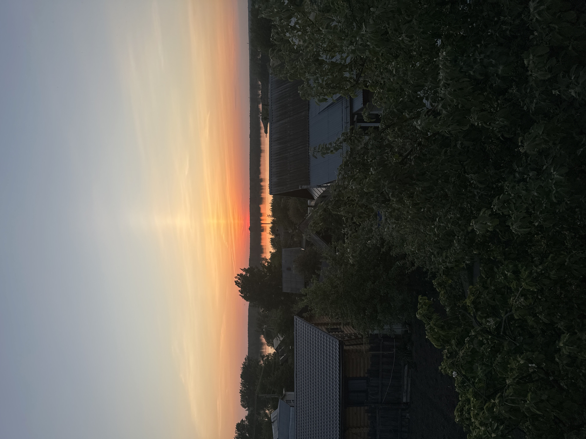 The image depicts a sunset over a body of water, likely a river or lake, with a village or town visible in the foreground. The sky transitions from light blue to orange and red hues, indicative of the setting sun. The water reflects the colors of the sky. Several buildings, likely residential homes, are visible, characterized by dark roofs and wooden structures. Trees are present, both near the buildings and along the shoreline. The overall impression is that of a tranquil, rural scene at dusk. The specific location as Chaikovsky, Russia is not definitively discernible.