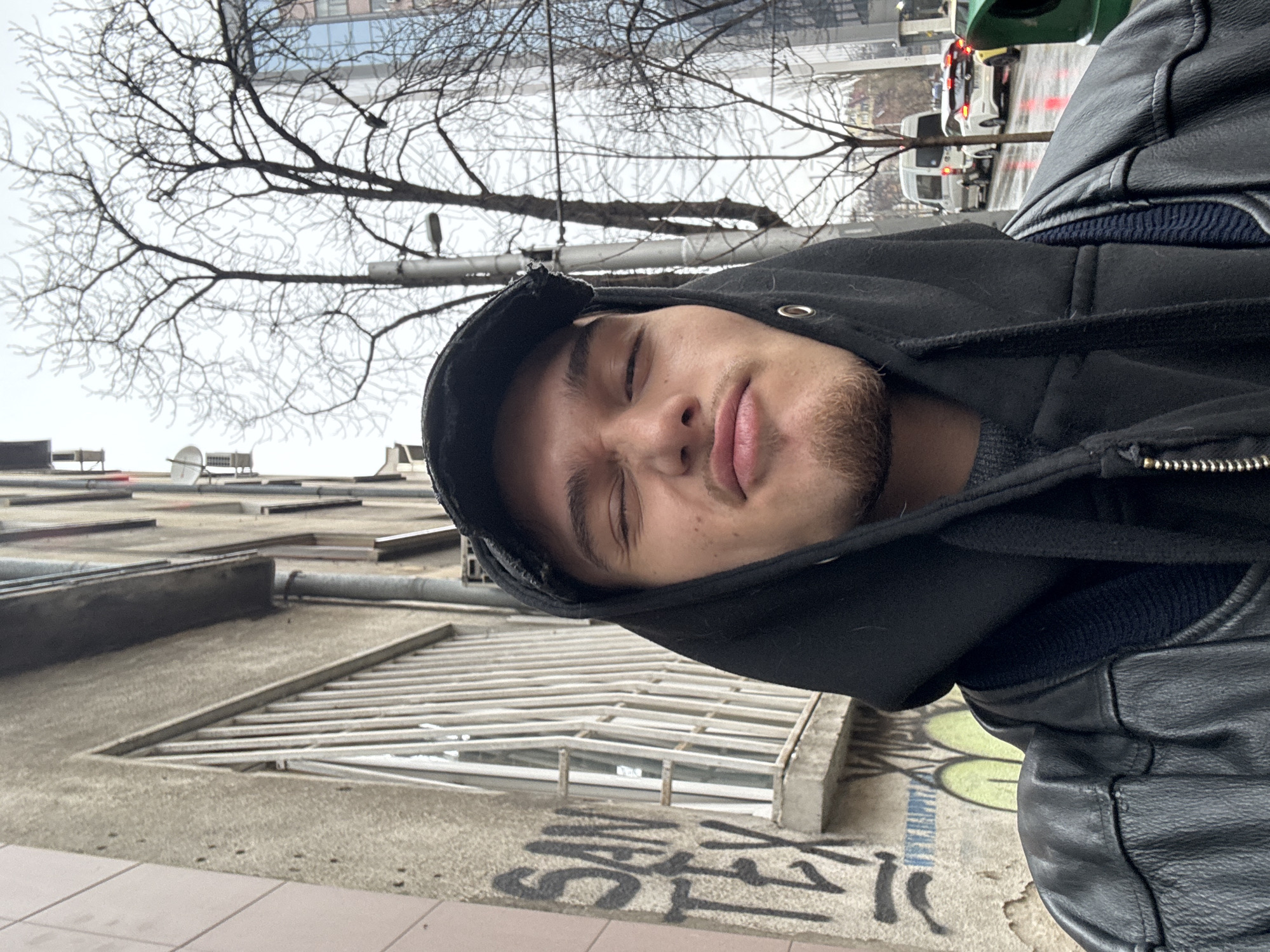 The photograph depicts a person wearing a black hooded jacket and a black leather jacket, likely taken in Sofia, Bulgaria. A gray building with grated windows and graffiti on the lower part of the exterior is visible. A bare tree branch extends into the upper part of the frame, suggesting a potentially cold, urban environment. In the background, blurred traffic and urban structures are observed, further supporting the Sofia location. The person's eyes are closed.