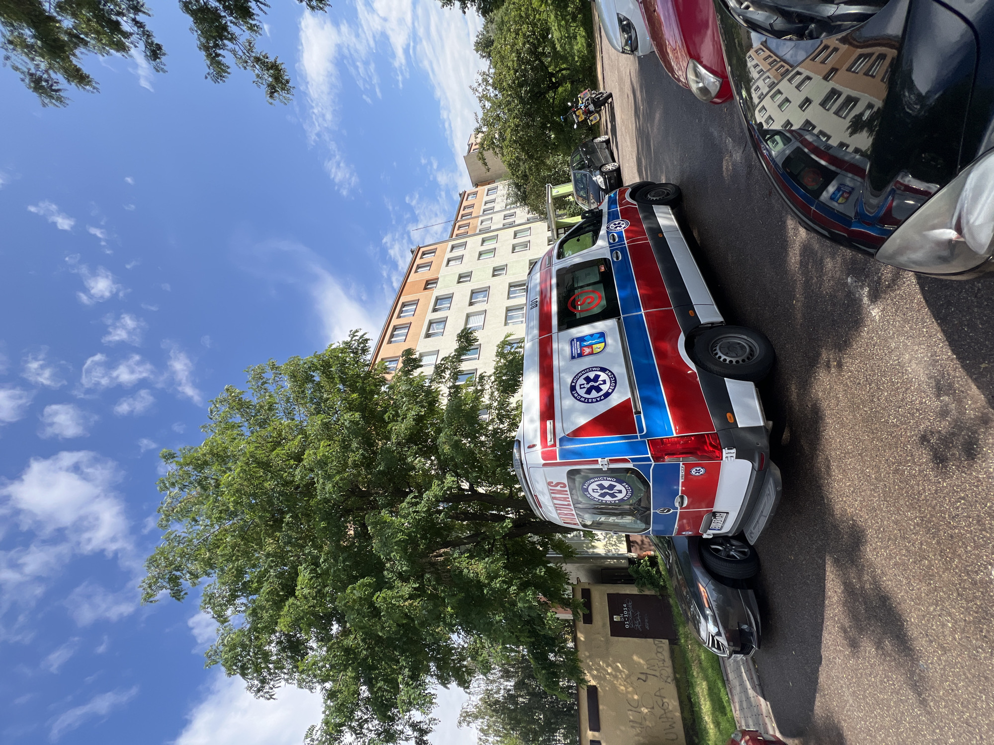 An ambulance is parked on a paved area in front of a multi-story apartment building. The ambulance is white with red and blue stripes and the word "AMBULANS" is visible on the side. There are several other vehicles parked nearby, including a dark sedan, a red car, and a motorcycle. The building appears to be residential, with multiple windows and balconies. Trees are visible in the background, and the sky is blue with some clouds. The scene appears to be a typical urban environment, likely in Białystok, Poland, given the markings on the ambulance.