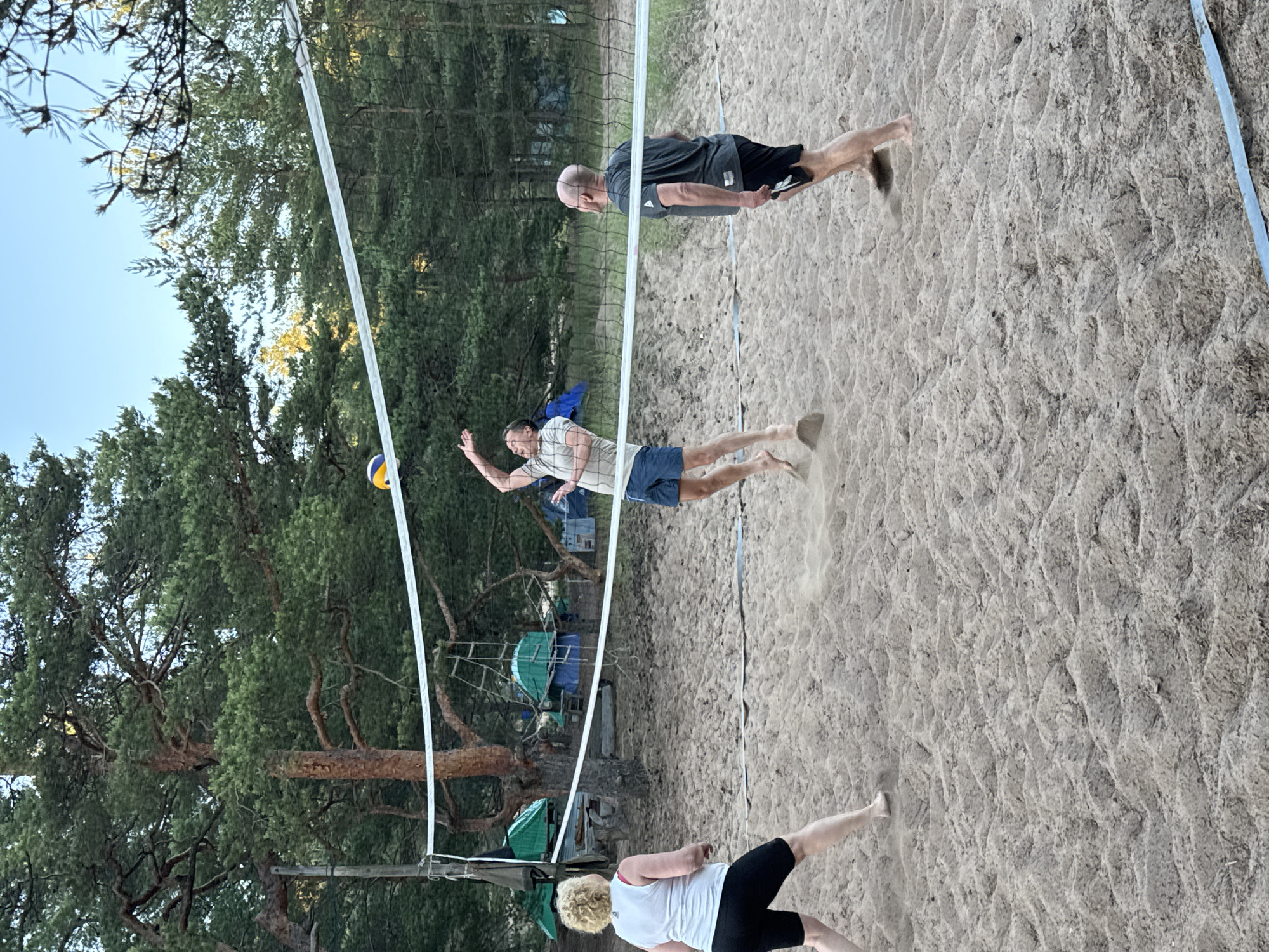 Three people are playing volleyball on a sandy court surrounded by pine trees. The court has a net stretched across it. In the foreground, a woman with blonde curly hair and a white tank top is facing away from the camera, wearing black shorts. To her right, a man in a light-colored t-shirt and blue shorts is mid-air, jumping to hit a volleyball that is in the air above him. To the right of the jumping man, a bald man with a dark t-shirt and black shorts is standing, facing the net. The background shows a forest with tall pine trees and some green tents are visible among the trees. The lighting suggests it is late afternoon or early evening.