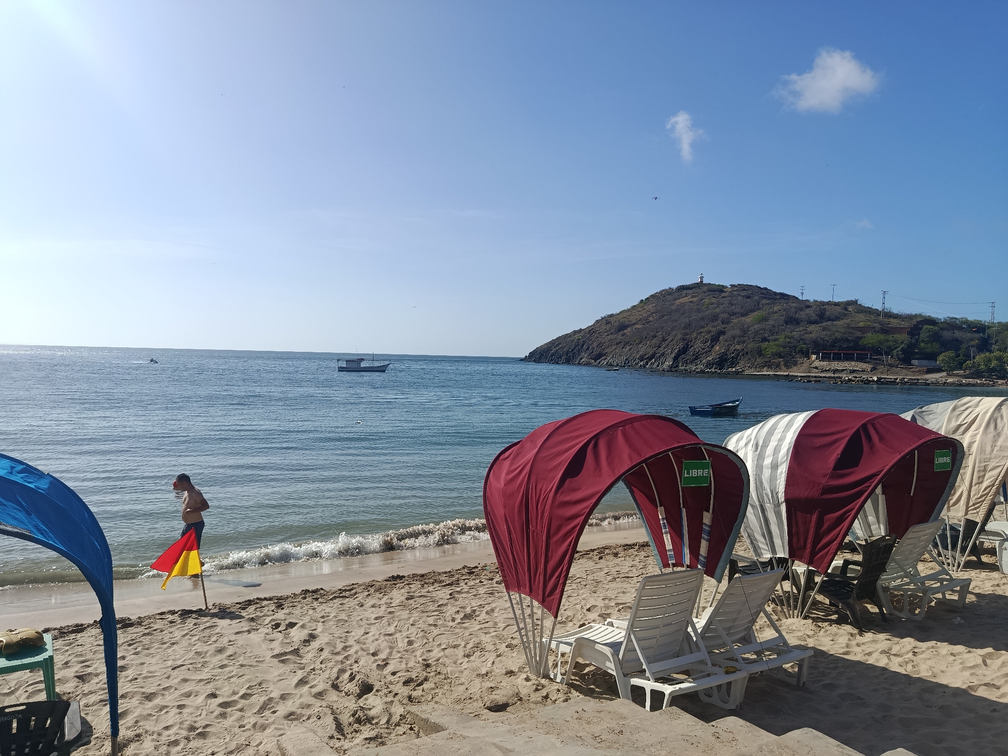 The image depicts a beach scene in Pedro González, Venezuela, under a clear blue sky. In the foreground, a sandy beach is lined with several beach chairs covered by awnings of varying colors, including maroon and striped red and white. A blue beach tent is partially visible on the far left. A person, appearing to be a young male, stands near the water's edge, holding a red and yellow flag attached to a pole. The ocean stretches out to the horizon, with a few boats visible on the water. In the background, a hilly, sparsely vegetated island with a lighthouse on its summit rises from the sea. The environment appears warm and sunny, characteristic of a tropical beach.