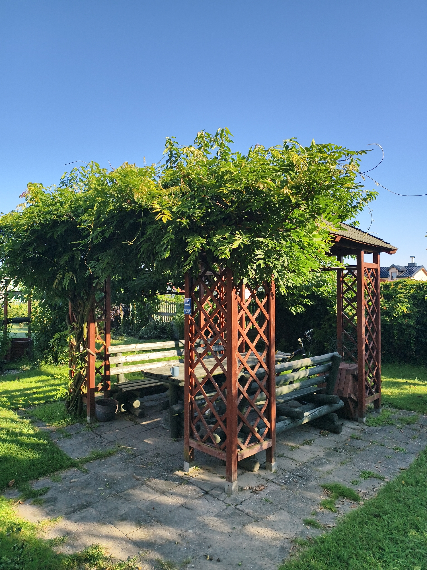 The image depicts a wooden gazebo covered with lush green foliage, providing shade over a picnic table and benches. The gazebo is situated outdoors in a grassy area with a paved pathway leading up to it. In the background, a green hedge and some houses are visible. The scene suggests a peaceful, outdoor setting, likely a garden or park. The location is identified as Tczew, Poland.