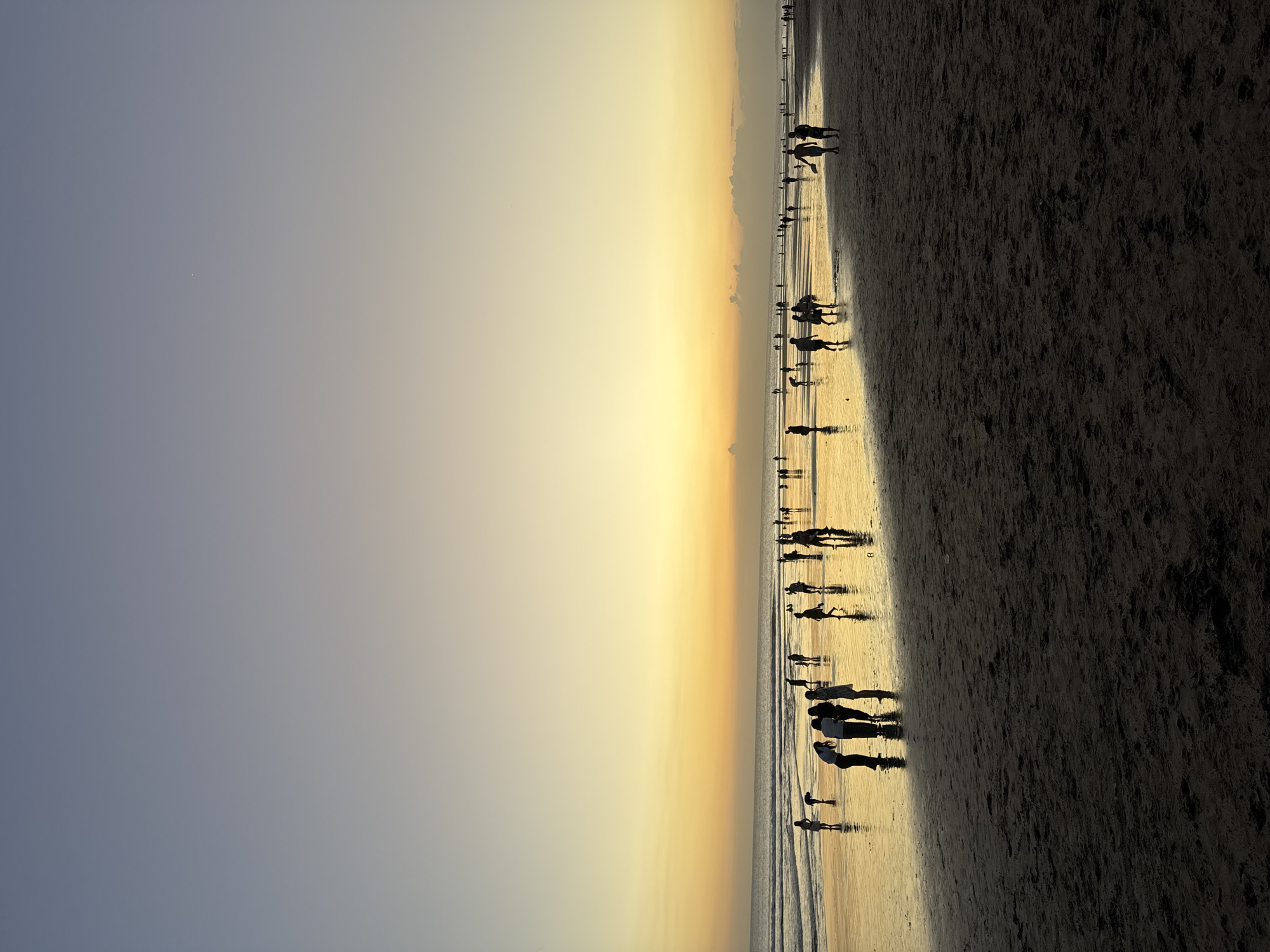 A wide shot captures the Kuta Beach in Bali, Indonesia, during a sunset. Silhouettes of people are spread across the wet sand, reflecting the warm hues of the sky. The ocean is calm, with gentle waves lapping at the shore. The sky transitions from a soft blue at the top to a vibrant yellow and orange near the horizon, creating a serene and picturesque atmosphere. A small dot, possibly a star or a planet, is visible in the upper portion of the sky. The beach is populated with individuals and small groups, some walking, others standing, with their figures casting elongated shadows on the wet sand. The scene is peaceful, with the setting sun casting a golden glow over the entire landscape.