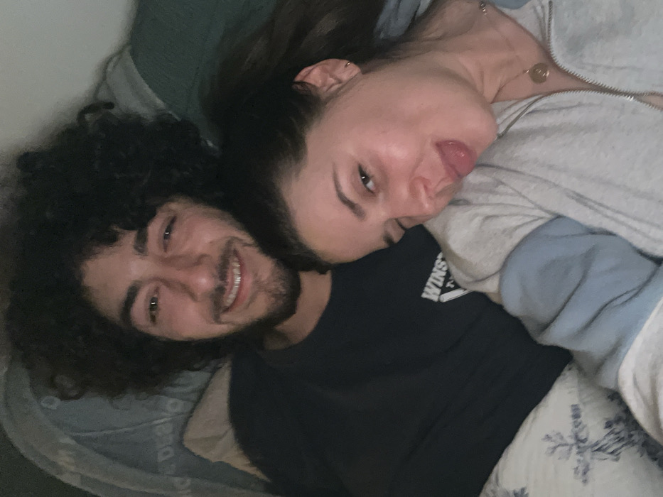 This image, located in Carnin, France, depicts two individuals in a dimly lit, indoor setting. A male subject, positioned on the left and slightly behind, exhibits dark, curly hair and a short beard. He wears a dark long-sleeved shirt with a white "WINSTON" graphic visible on the chest, and his mouth is open in a smile. A female subject, positioned on the right and slightly in front, has dark hair pulled back into a ponytail. Her left eye is closed in a wink, and her tongue is extended. She wears a light-colored zip-up hoodie with light blue accents on the sleeves and a gold-toned necklace with a circular pendant. The subjects' heads are in close proximity. The background includes a light-colored pillow with a blue foliate pattern on the left, and a darker, indistinct fabric object on the right.