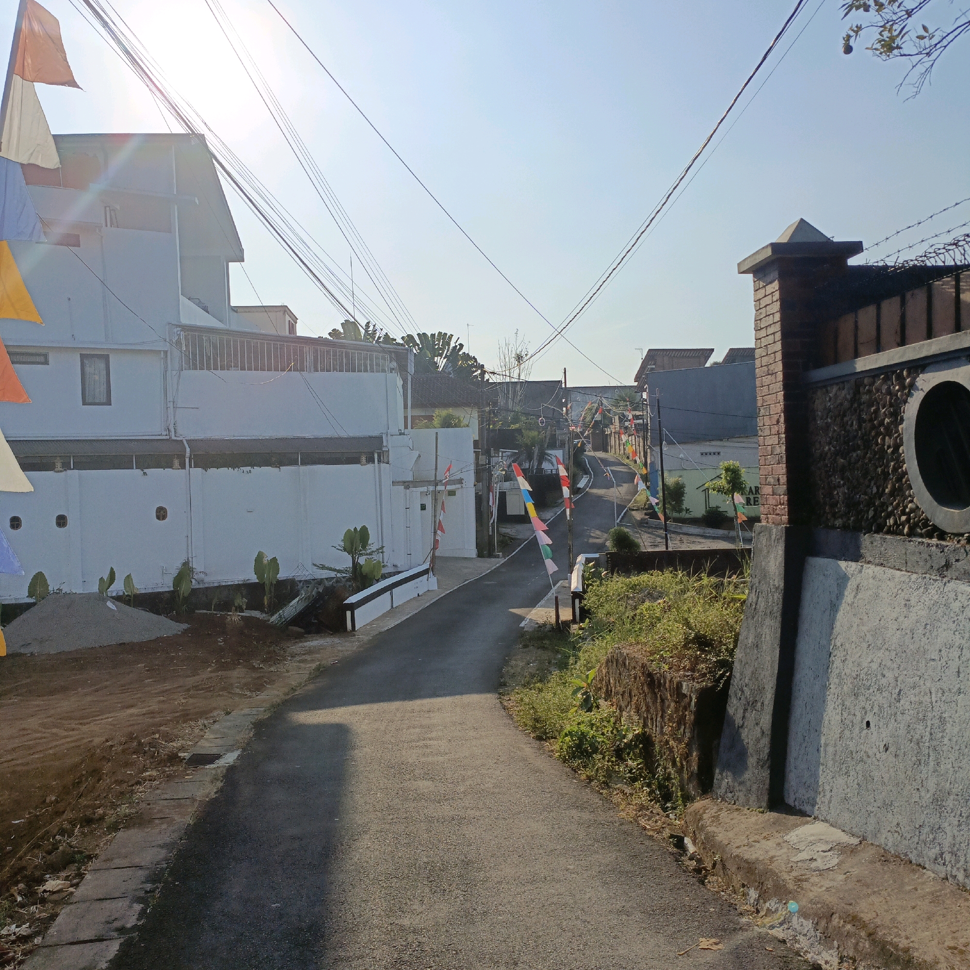 This image depicts a narrow, paved road winding through a residential area. The road is lined with houses and fences. Small flags on poles, in red, white, blue, and yellow, are strung along the street, suggesting a festive occasion or holiday. The buildings are modern, mostly painted white, with some featuring ornate brickwork on fences and pillars. Vegetation, including bushes and trees, is visible along the roadside and in front of the houses. The sky is clear blue, with some power lines crisscrossing overhead. The scene is illuminated by bright sunlight, casting shadows on the road. The overall impression is a quiet, sunny day in a well-kept, possibly suburban, neighborhood in Salatiga, Indonesia.