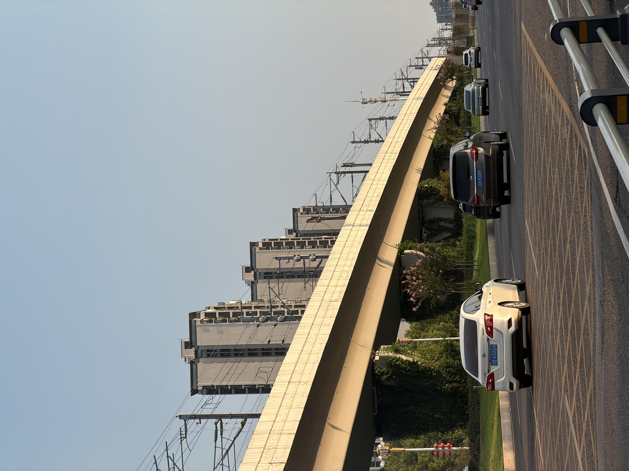 The image depicts a street scene in Jiangjin, China, during what appears to be late afternoon or early evening, indicated by the warm, angled sunlight. The foreground shows a multi-lane road with yellow lane markings and pedestrian crossing patterns. Several vehicles are on the road: a white sedan with license plate "SB 63866" in the left lane, a dark SUV with license plate "A 9C009" in the center lane, a silver sedan, and a white SUV further ahead. Overhead, a raised concrete overpass, likely for a train or elevated roadway, stretches across the scene, supported by columns partially covered in greenery. The overpass is lined with power lines and poles. In the background, a series of modern residential buildings with a light gray facade are visible, aligned along the overpass. The sky is clear and pale blue. Greenery, including trees and bushes, lines the sides of the road and the base of the overpass. A street light with red signals is visible on the far left.