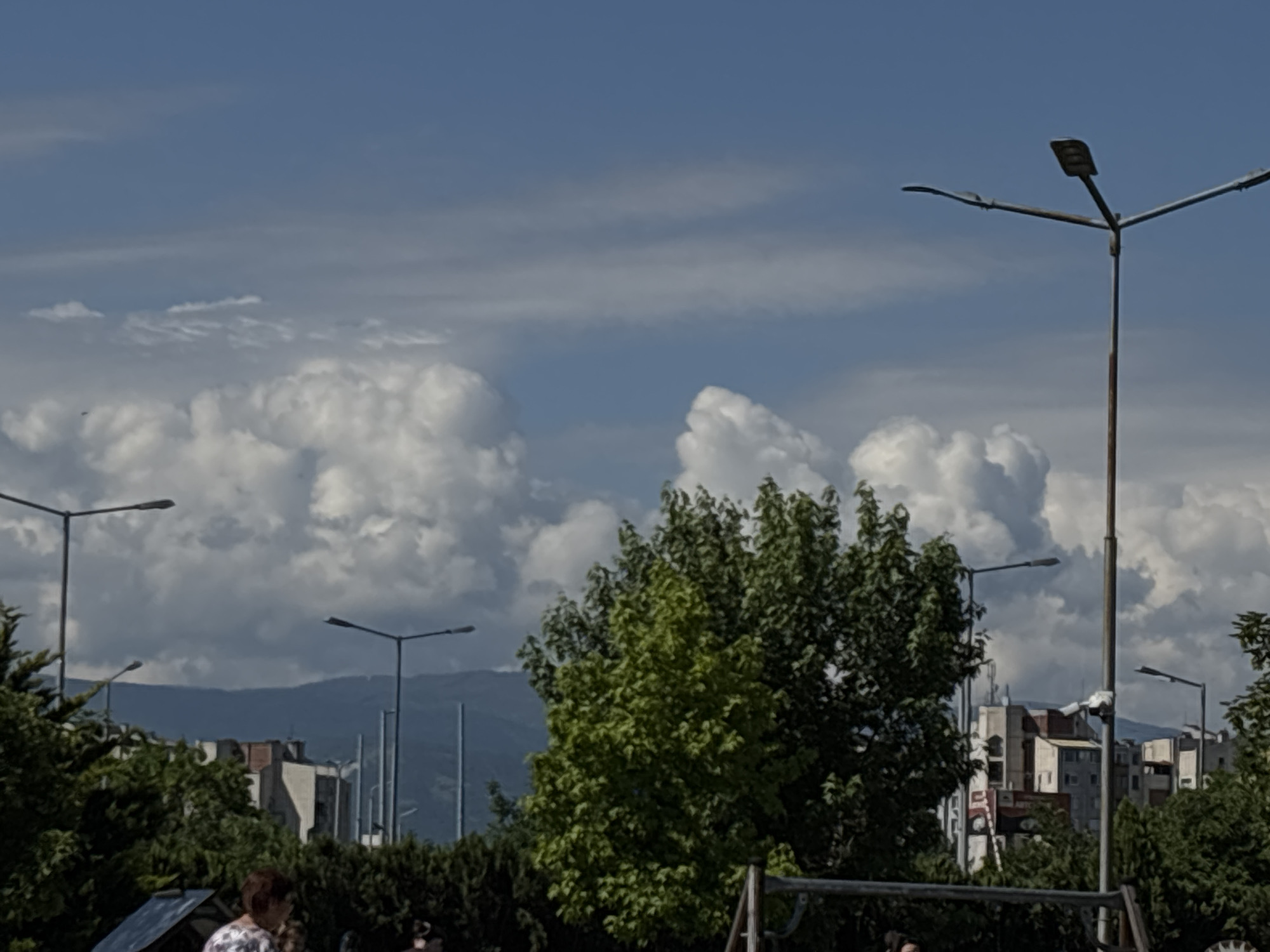 The image depicts an outdoor scene in Plovdiv, Bulgaria. In the foreground, there is green vegetation, including a tree with both light and dark green foliage. Behind the vegetation are several buildings with different heights, some appearing to be residential apartments. There are street lamps with horizontal arms and lights present. The sky is light blue with large, white, puffy clouds. In the midground, there is a hint of a mountain range in the distance. A possible playground structure is present at the bottom of the image. A person is partially visible on the left.
