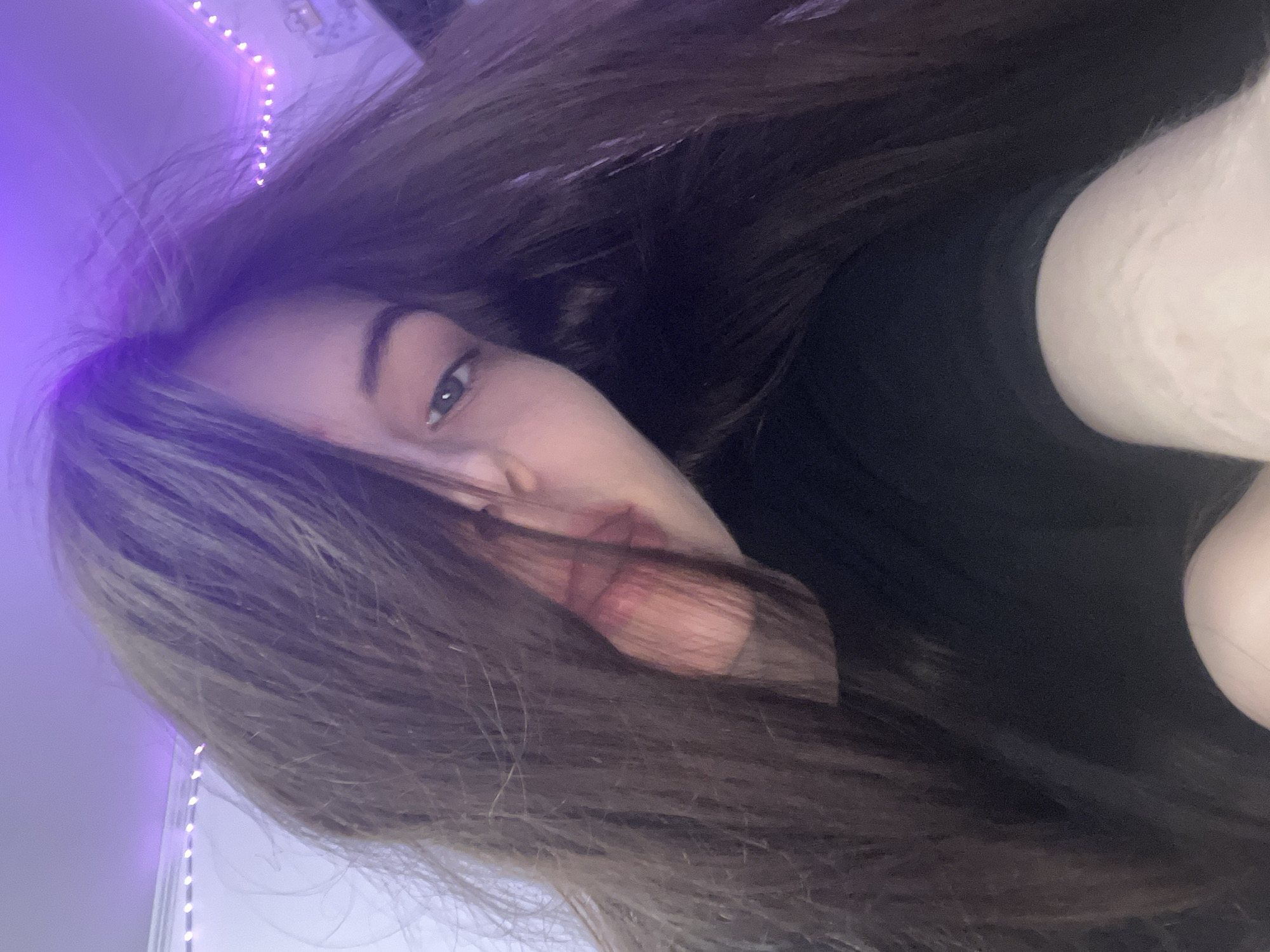 The image shows a young person with long, brown hair partially covering their face. They have fair skin and one eye is visible, appearing to be blue or gray. The person is wearing a black top. The background is blurred and includes a wall with purple LED lights. The location is not discernible from the image. There is nothing in the image to indicate that the image was taken in Saint Petersburg, Russia.