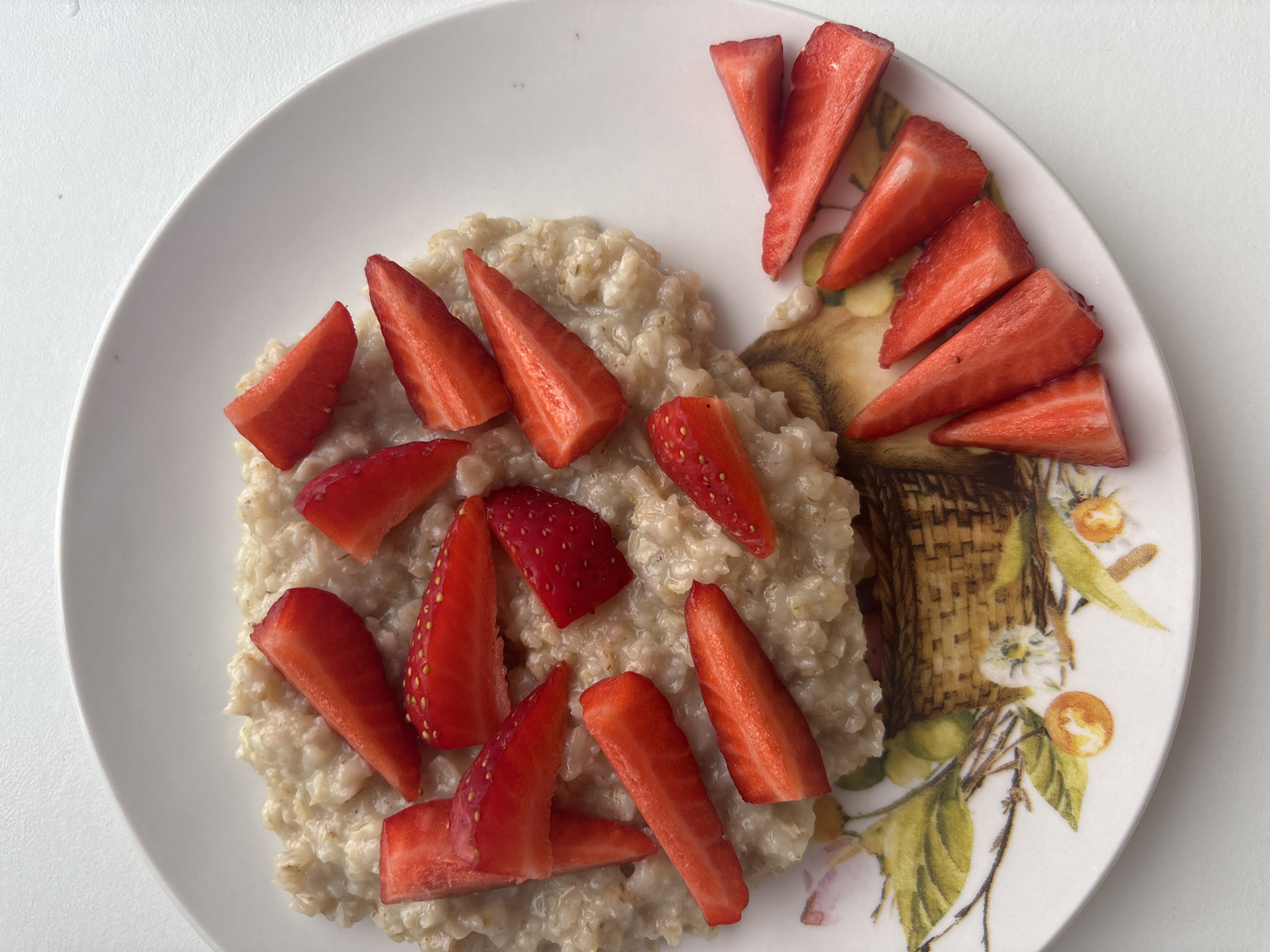 A plate of oatmeal topped with sliced strawberries sits on a white surface. The oatmeal is light brown in color and appears to be slightly watery. The strawberries are bright red and are cut into triangular wedges. Some strawberry slices are placed directly on top of the oatmeal, while others are arranged around the edge of the plate. The plate itself is white with a floral design featuring a basket and foliage around its rim. The location is likely Chaikovsky, Russia, based on external data, but is not evident in the image itself.