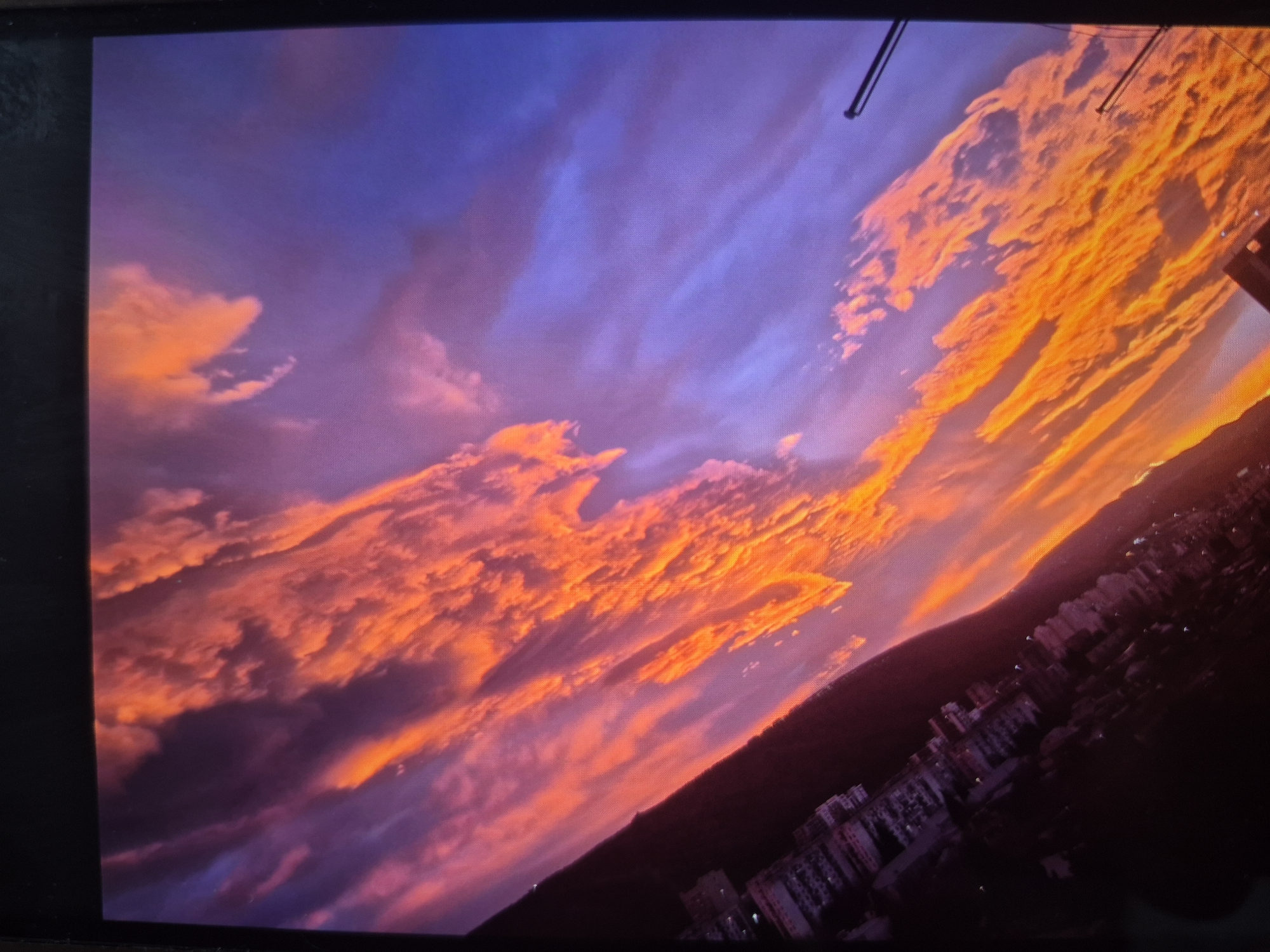 The image shows a vibrant sunset over Tbilisi, Georgia. The sky is a mix of blue and purple hues transitioning into orange and red near the horizon. The clouds are layered and illuminated by the setting sun, creating a dramatic effect. Below the horizon, there is a dark mass, which is likely a mountain range, with urban structures at its base including densely-packed buildings, suggesting a city landscape. The scene suggests a high vantage point, possibly from a high-rise building or a hill overlooking the city. There is a visible cable or wire in the upper right corner of the image, indicating the possible location of the picture.