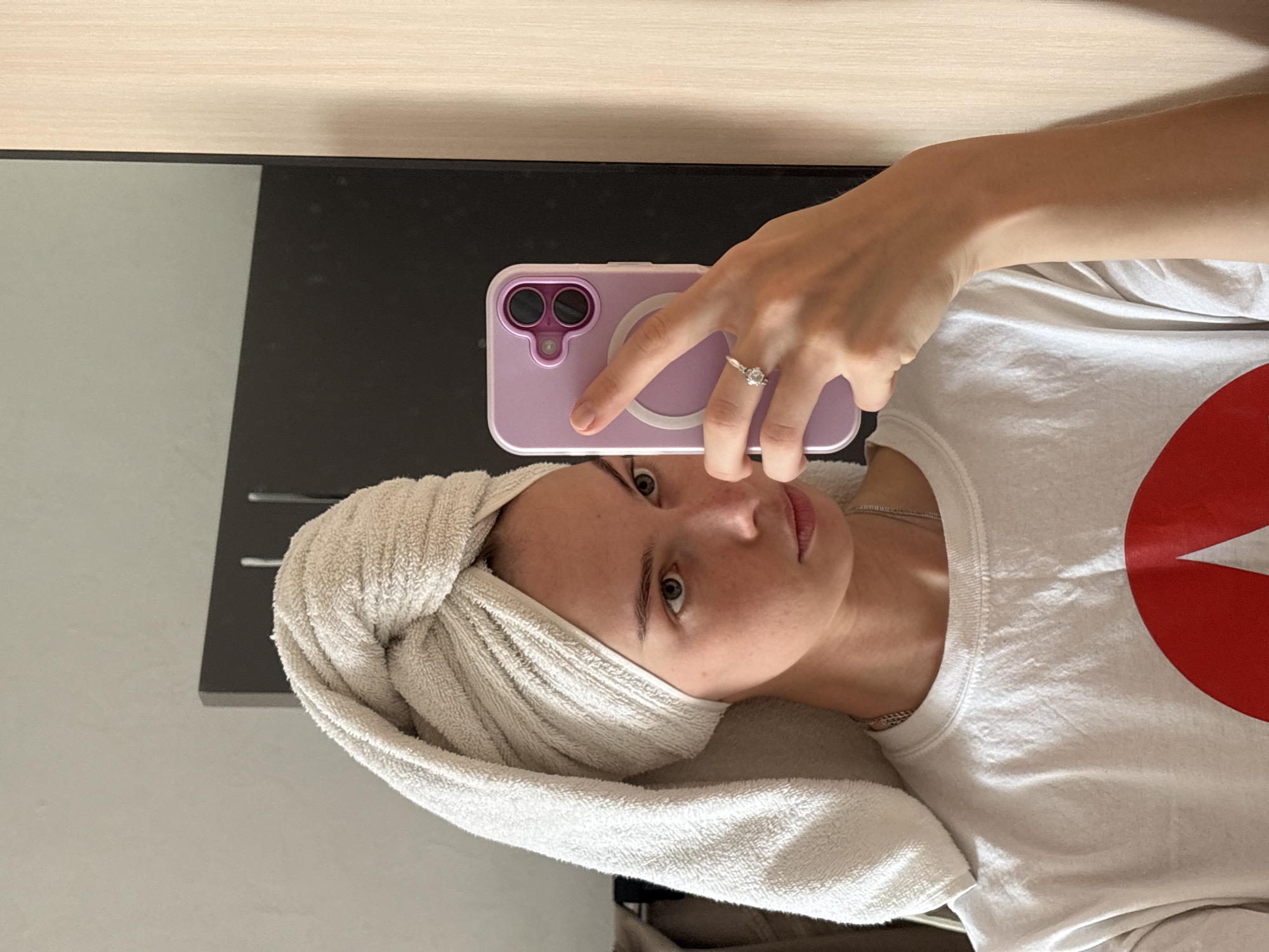 A young woman with a towel wrapped around her head is taking a selfie in a mirror. She is wearing a white t-shirt with a red graphic on it and a silver necklace. She is also wearing a ring on her left hand. The background appears to be a room with a mirror and a closet. The image is taken from a slightly elevated angle. The location in the city of Perm, Russia is not discernible from the image.