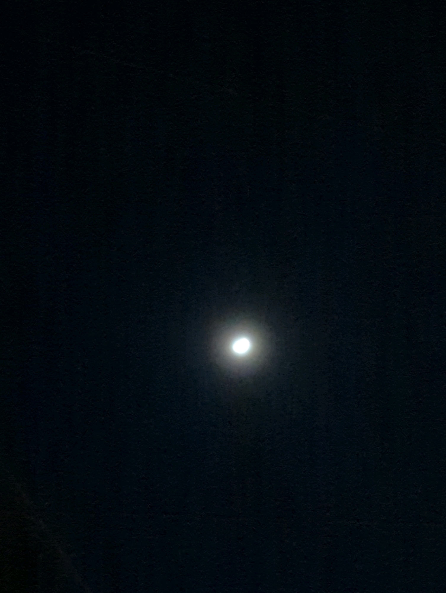 A full moon is visible in a dark night sky. The moon is located in the center of the frame and is illuminated by an external light source, suggesting it may be a streetlamp or other artificial light reflecting off the moon's surface. The background is a dark, textured surface, possibly a wall or ceiling. There is no indication of the specific location within Musanze District, Rwanda, as the image focuses solely on the celestial body. No people, subjects, objects, or actions are depicted other than the moon itself.