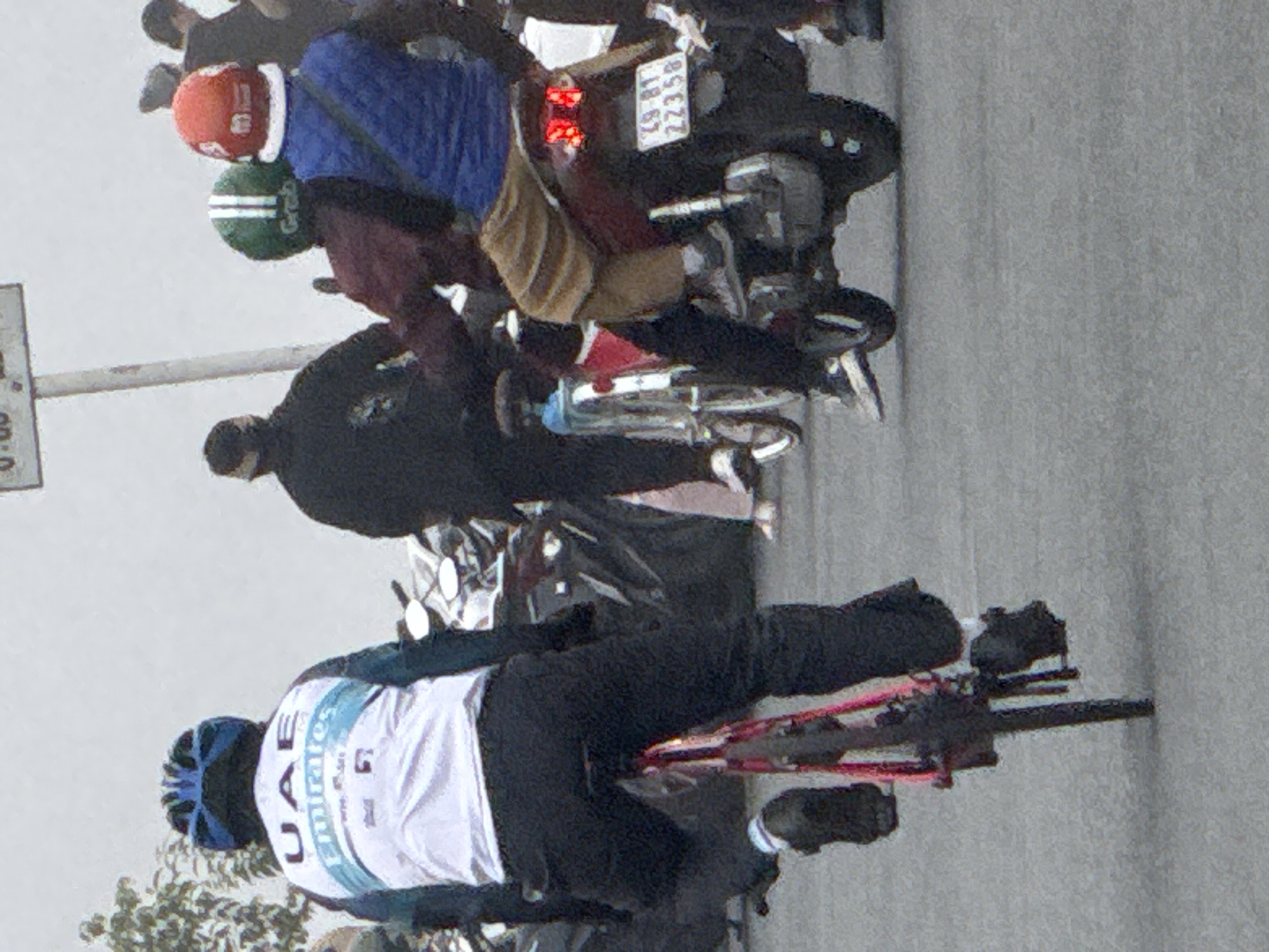 A group of people are gathered around motorcycles and bicycles on a paved road in Da Lat, Vietnam. The foreground features a person on a bicycle wearing a blue and white cycling jersey with "UAE Emirates" printed on it and a blue helmet. They are wearing black pants and black shoes with white socks. To the right, two people are on a motorcycle. The person in the front is wearing a red helmet and a blue vest over a jacket. The person behind them is wearing a green and white helmet and a maroon jacket. The motorcycle has a license plate that reads "49-B1 22358". In the background, other people are visible near more motorcycles, and trees line the left side of the frame. The sky is overcast.