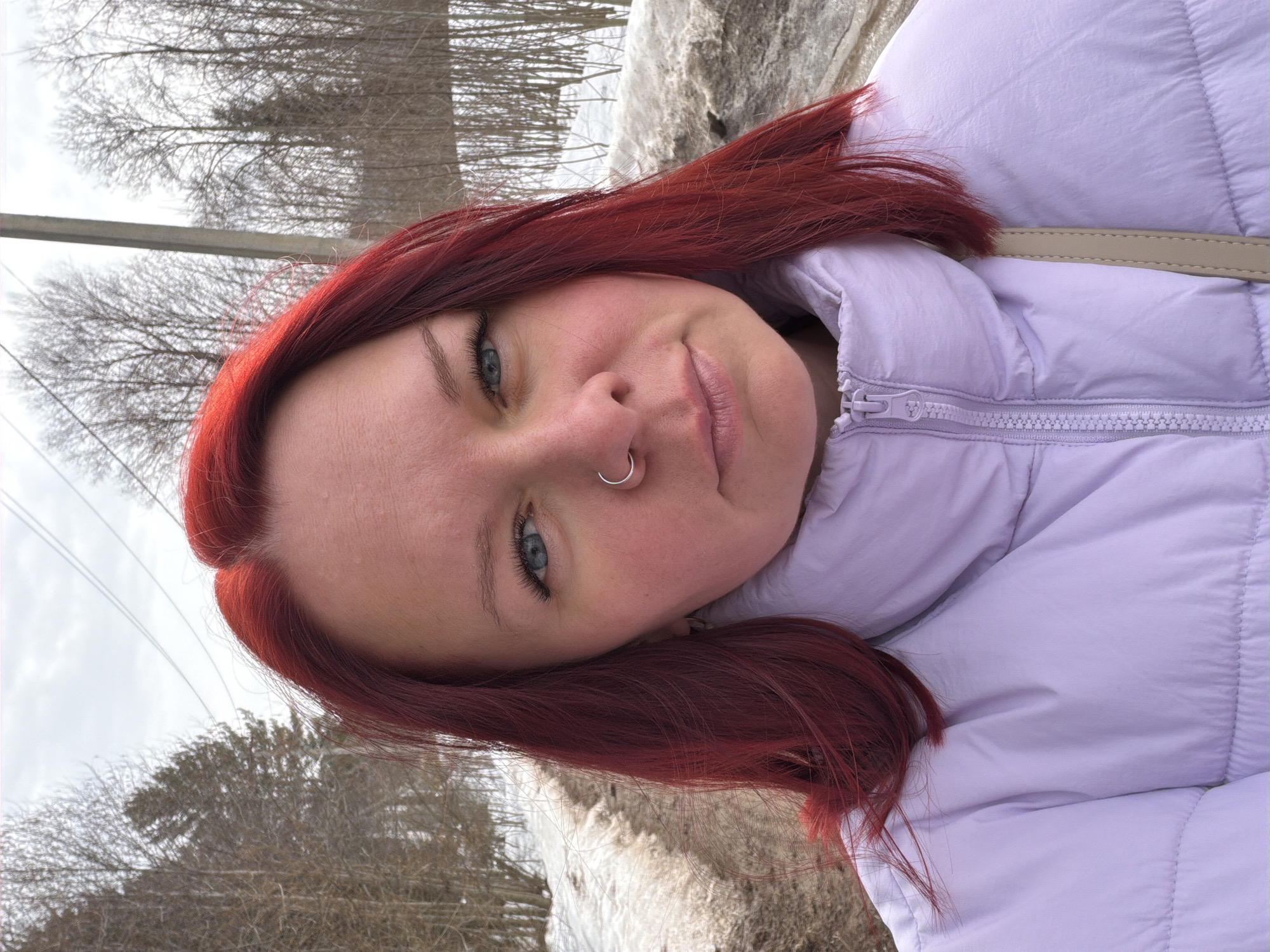 The image presents an outdoor, mid-shot portrait of a woman located in Chaikovsky, Russia, during what appears to be late winter or early spring. She has long, vibrant red hair and blue eyes, with a silver-colored hoop piercing visible in her left nostril. Her complexion is fair, with some redness on her cheeks. She wears a light lavender (light purple) quilted puffer jacket, zipped up, and a beige strap, likely from a bag, is visible over her right shoulder. Her gaze is directed forward, directly at the camera, with a neutral expression.
The background shows a partially snow-covered dirt or unpaved road receding into the distance on the left side of the frame. The ground exhibits patches of melting snow and exposed earth. A dense line of bare deciduous trees and some coniferous trees forms a forest on both sides of the path. An overcast, light grey sky is visible above the treeline. A grey utility pole and associated power lines are positioned vertically on the right side of the background.