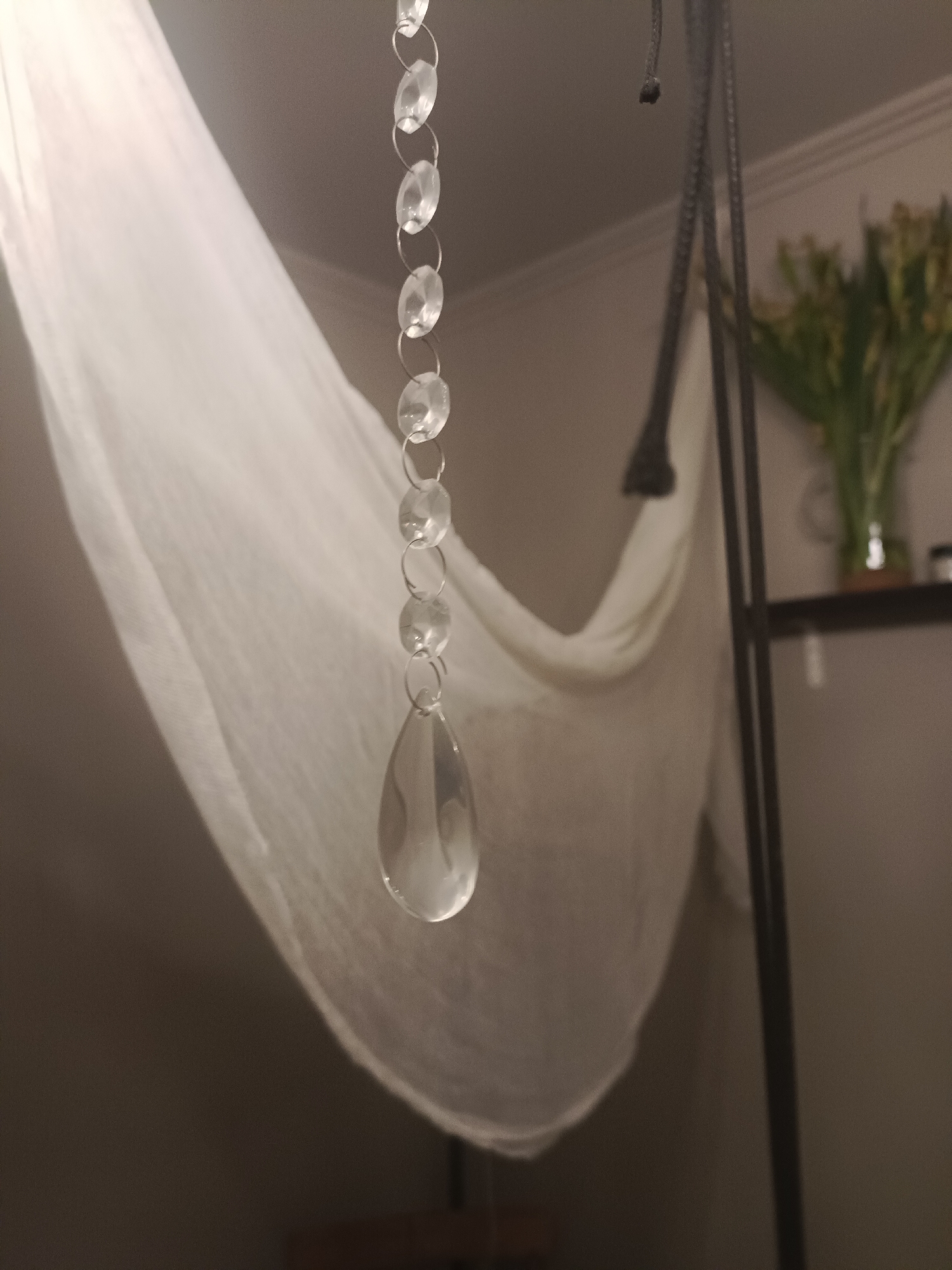 The image captures a close-up of a string of clear faceted beads, likely crystal or glass, hanging vertically. The beads are strung together with small silver rings. The largest bead at the bottom is teardrop-shaped and shows significant light refraction. In the background, a sheer white fabric, possibly a curtain or draped material, fills the left side of the frame, creating soft folds and shadows. To the right, a dark vertical pole is visible, part of a stand or structure, and behind that, a shelf holds a glass vase with yellow flowers. The lighting is dim, with the primary light source appearing to illuminate the beads from above and the left, causing reflections and highlights. The overall scene suggests an indoor setting, possibly a home or a studio. The image does not contain any elements that would place it specifically in Moscow, Russia.