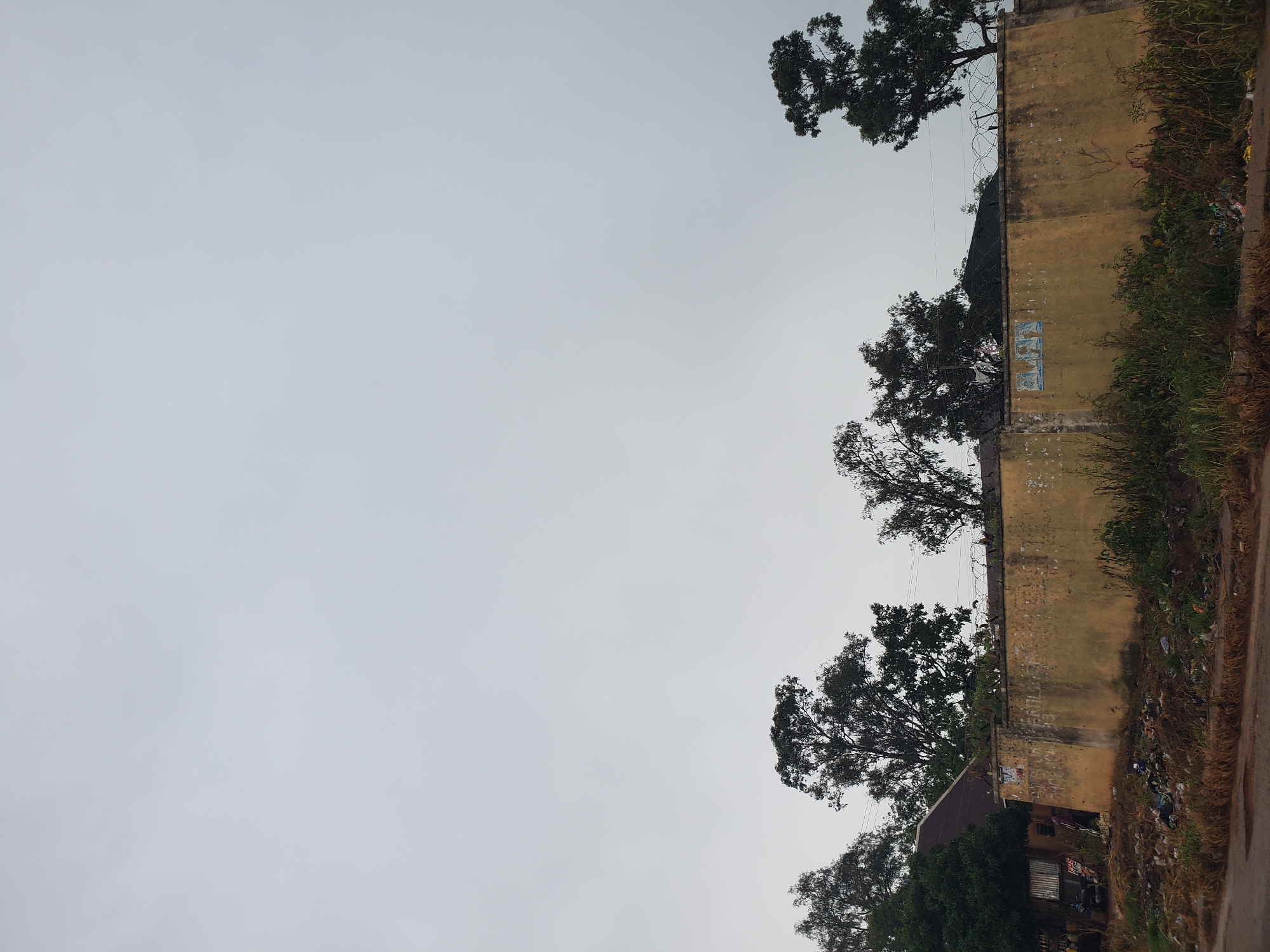 The image depicts a wall of a building in Barikin Gidar Kanat, Nigeria, under an overcast sky. The wall, painted a faded yellow, appears aged and weathered, with some peeling paint and scuff marks. Barbed wire is stretched across the top of the wall. Several trees with lush green foliage are visible, some reaching over the wall and others in the foreground. One section of the wall has three faded rectangular posters or signs adhered to it. At the base of the wall, there is a pile of debris and trash, with some vegetation growing around it. The overall scene suggests an outdoor environment, possibly a residential or commercial area, with signs of neglect and an accumulation of waste. The time of day appears to be daytime, indicated by the natural light, and the weather is cloudy. There is no clear activity or event taking place; the image seems to be a still, observational shot of the setting. The image is oriented vertically, with the sky occupying the majority of the left side and the wall and trees on the right.