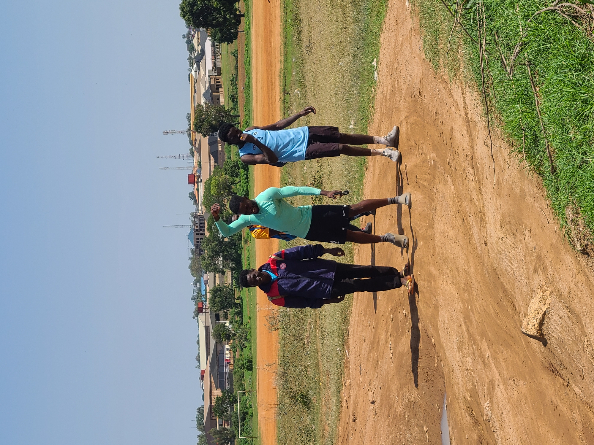 In Barikin Jirga Summa, Nigeria, three young men are captured mid-stride on a dusty, dirt path. The man on the left, wearing a blue, red, and white jacket and black trousers, smiles at the camera. The central figure, clad in a teal long-sleeved athletic shirt and black shorts, strikes a pose with his arm raised. To the right, a man in a blue tank top and dark shorts, with a backpack, appears to be eating something. The background reveals a rural or semi-urban landscape with a large, orange, dusty field, possibly a sports field, bordered by green grass and trees. Beyond that, buildings and communication towers are visible under a clear blue sky.