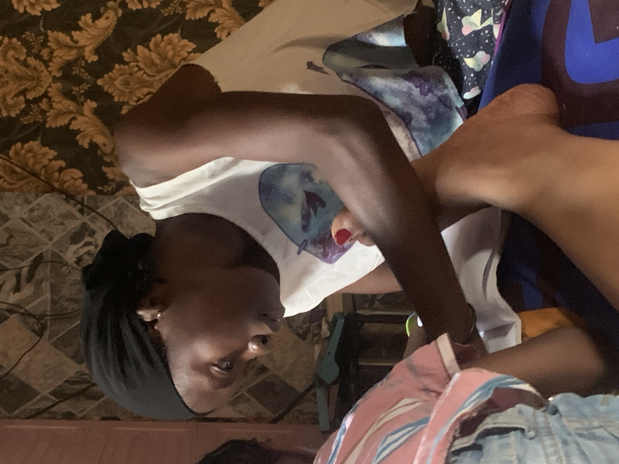 The media file shows a close-up, rotated view of a person, likely a young woman, lying down on a patterned couch. She is wearing a white t-shirt with a circular graphic and a black headscarf. Her head is resting on a patterned blanket or cushion, and her arm is bent at the elbow. Another person's leg, with painted red toenails, is visible in the foreground, suggesting they are also on the couch or nearby. The background features a dark, floral-patterned fabric, possibly part of the couch or wall covering, and a floor with a tiled or patterned surface. The lighting is dim, suggesting an indoor setting, possibly in the evening or with artificial light. There is no visible text in the image. The scene appears casual and relaxed, with the subjects in a reclining position. The overall impression is of a personal, candid moment captured in a domestic environment within Sabon Tasha, Nigeria.