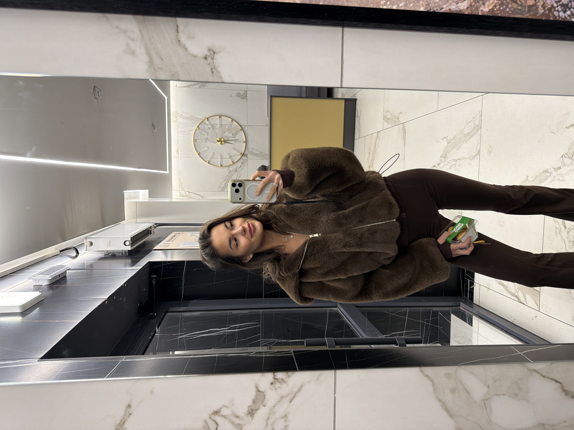 A young woman stands in a bathroom, taking a selfie in the mirror. She is wearing a brown fur jacket and matching pants. She is holding a phone in her left hand and a package of cookies in her right hand. The bathroom has marble walls and countertops, a modern sink and faucet, and a clock on the wall. Based on the clock and marble tile work in the bathroom, and the person holding cookies, it is likely that the location is a hotel in the city of Krasnoyarsk, Russia.