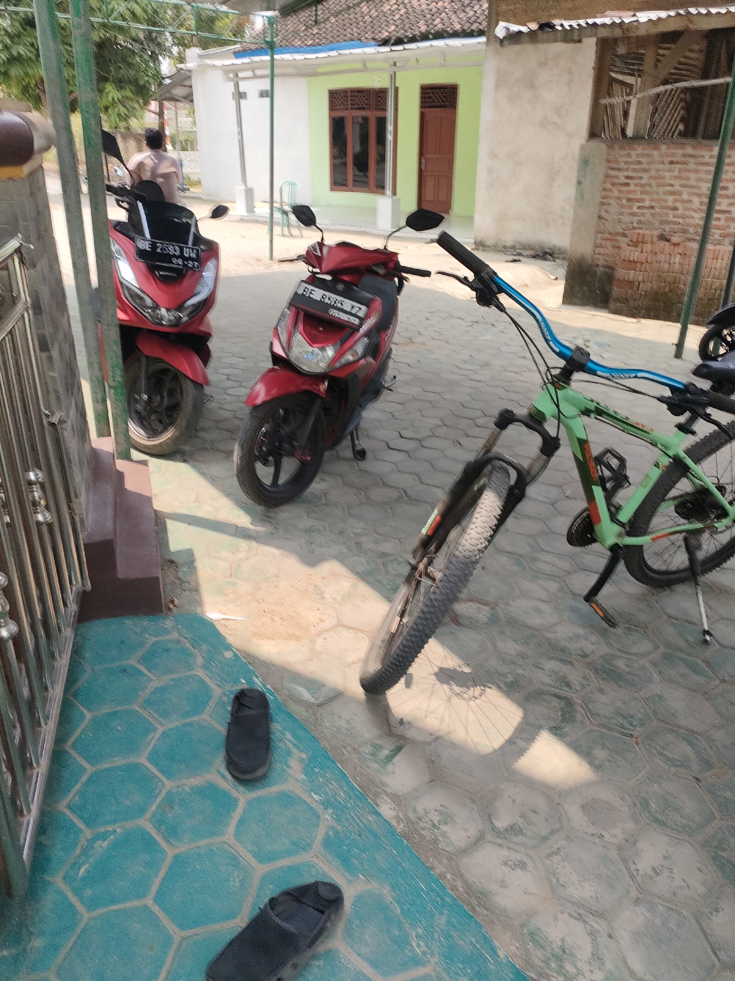 Two red scooters and a light green bicycle are parked on a paved outdoor area with hexagonal tiles in Pringsewu Barat, Indonesia. A person in a light-colored shirt is walking away from the scooters in the background. The scooters are positioned to the left of the frame, one slightly in front of the other. The bicycle is to the right, with its handlebars turned towards the viewer. A metal railing and steps are visible in the lower left corner. The environment appears to be a residential or commercial area with buildings and a fence. The ground is a combination of hexagonal tiles and concrete.