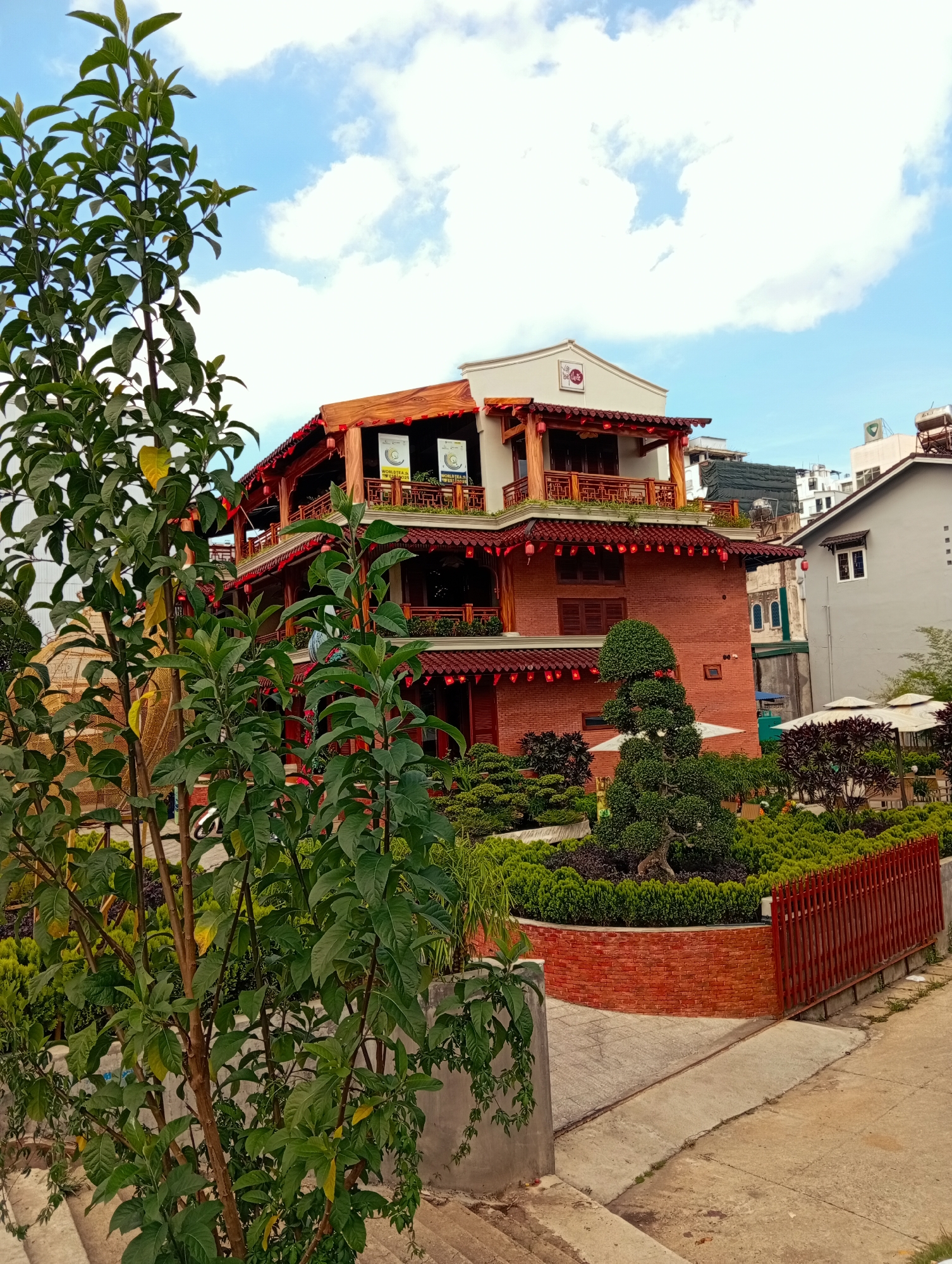 The image presents a striking multi-story building with distinct East Asian-inspired architecture, set against a clear blue sky dotted with white clouds. The building features prominent red tiled roofs, intricately carved wooden railings, and multiple balconies adorned with numerous small red lanterns, creating a vibrant aesthetic. Its lower section is constructed from red brick, complementing the red accents of the upper wooden structures and white facade.
In the foreground, a large, leafy green tree partially obscures the left side of the building, adding a natural, lush element to the scene. The building is surrounded by a meticulously maintained garden area, featuring sculpted green bushes, dark foliage plants, and topiaries, including one prominently shaped like a tiered tree. White outdoor umbrellas are visible within the garden, hinting at outdoor seating.
A low red brick wall and a matching red metal fence define the perimeter of the garden space along a concrete path and steps in the foreground. In the background, other urban buildings are visible, suggesting an integrated urban environment.
Visible text includes a logo on the top white facade of the building, appearing to read "Red Cafe" or similar, stylized with a heart shape. Additionally, banners hanging from the upper balcony clearly display "WORLD TEA & COFFEE EXPO 2023," indicating the building's commercial nature, likely a tea house or cafe, or its association with such an event.
The scene depicts a bright, clear day, suggesting good weather. No people are visible, and no specific activity is captured, presenting a tranquil and inviting view of the establishment and its surrounding landscape, consistent with the unique charm often found in locations like Da Lat, Vietnam.