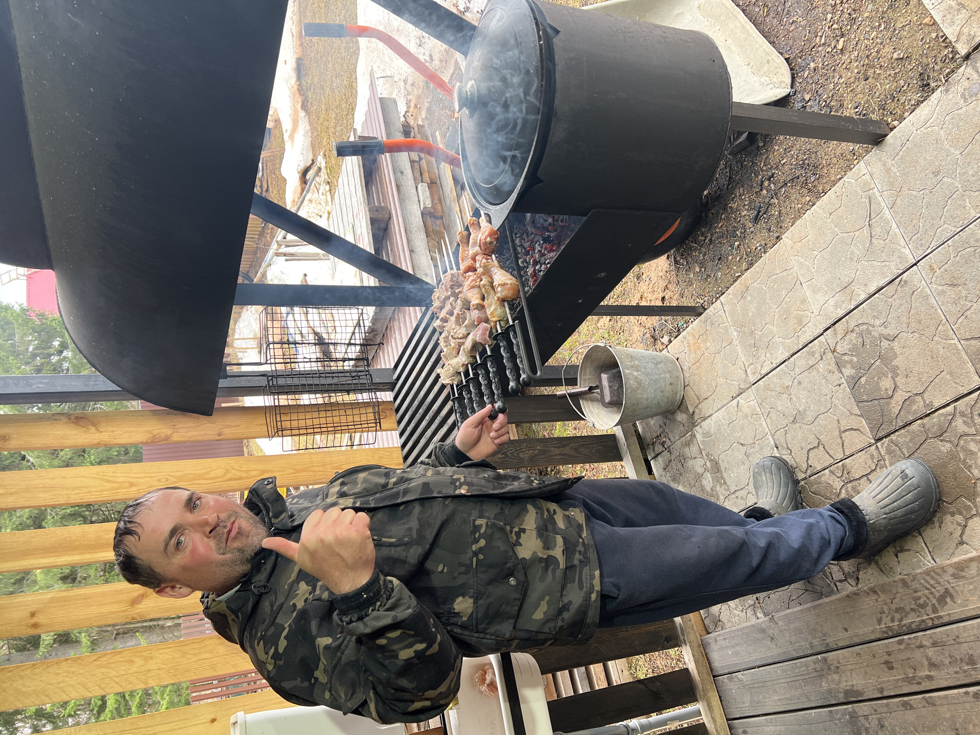 A man stands on a wooden deck beside a black, barrel-shaped barbecue grill. He wears a camouflage jacket, dark pants, and gray boots. He gestures with a thumbs-up. Skewers of meat are cooking on the grill. The environment includes wooden structures and ground covered with paving stones and vegetation. The location may be Bol'shakino, Russia.