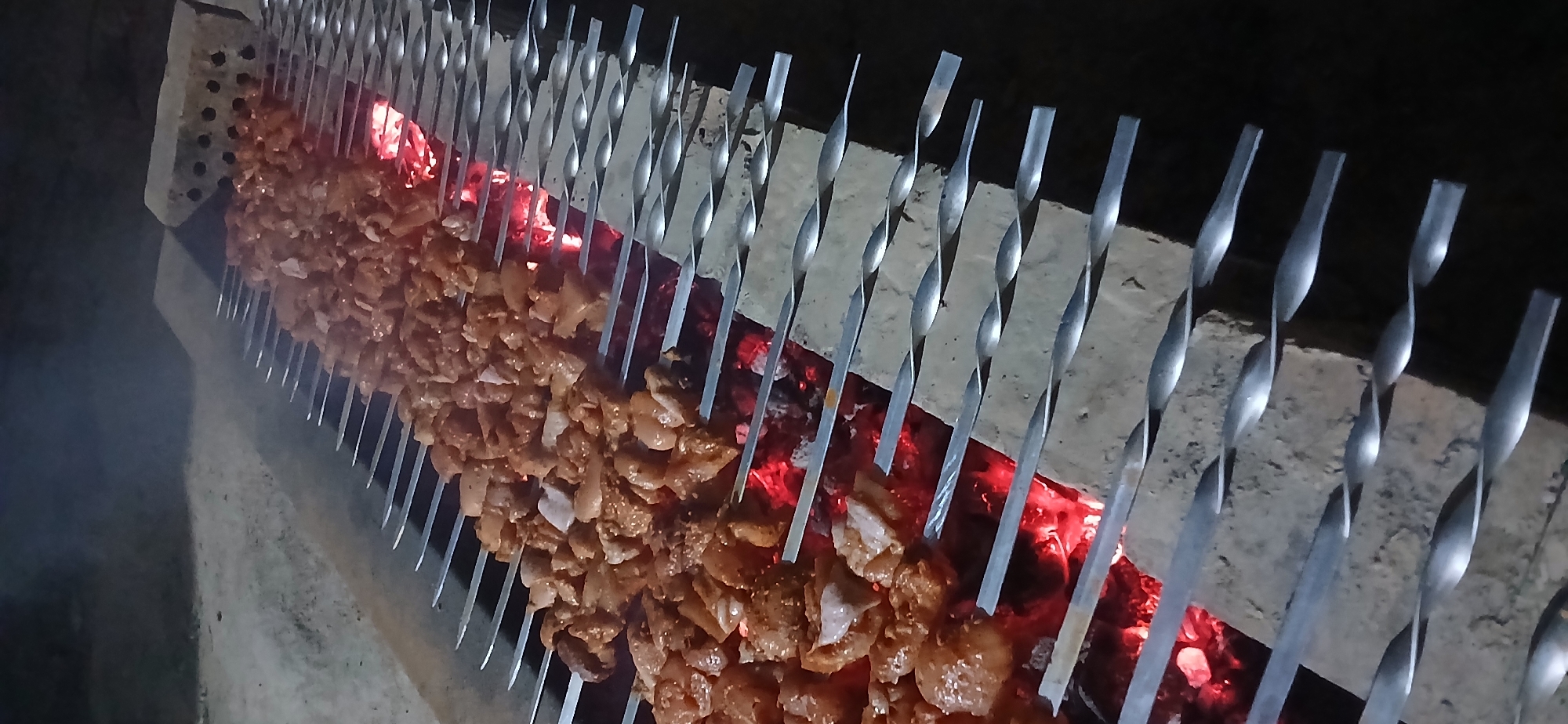 In Daliguzar, Uzbekistan, skewers laden with seasoned meat are being grilled over glowing red coals. The meat, cut into uniform pieces, is arranged densely on long metal skewers. The skewers are supported by a structure made of bricks, positioned over an outdoor fire pit. Smoke rises from the coals and the grilling meat, indicating the active cooking process. The scene is illuminated by the fire's glow, suggesting it is nighttime or a dimly lit area. The background is dark and indistinct, with some sandy or dusty ground visible. The overall impression is of a traditional outdoor barbecue.
