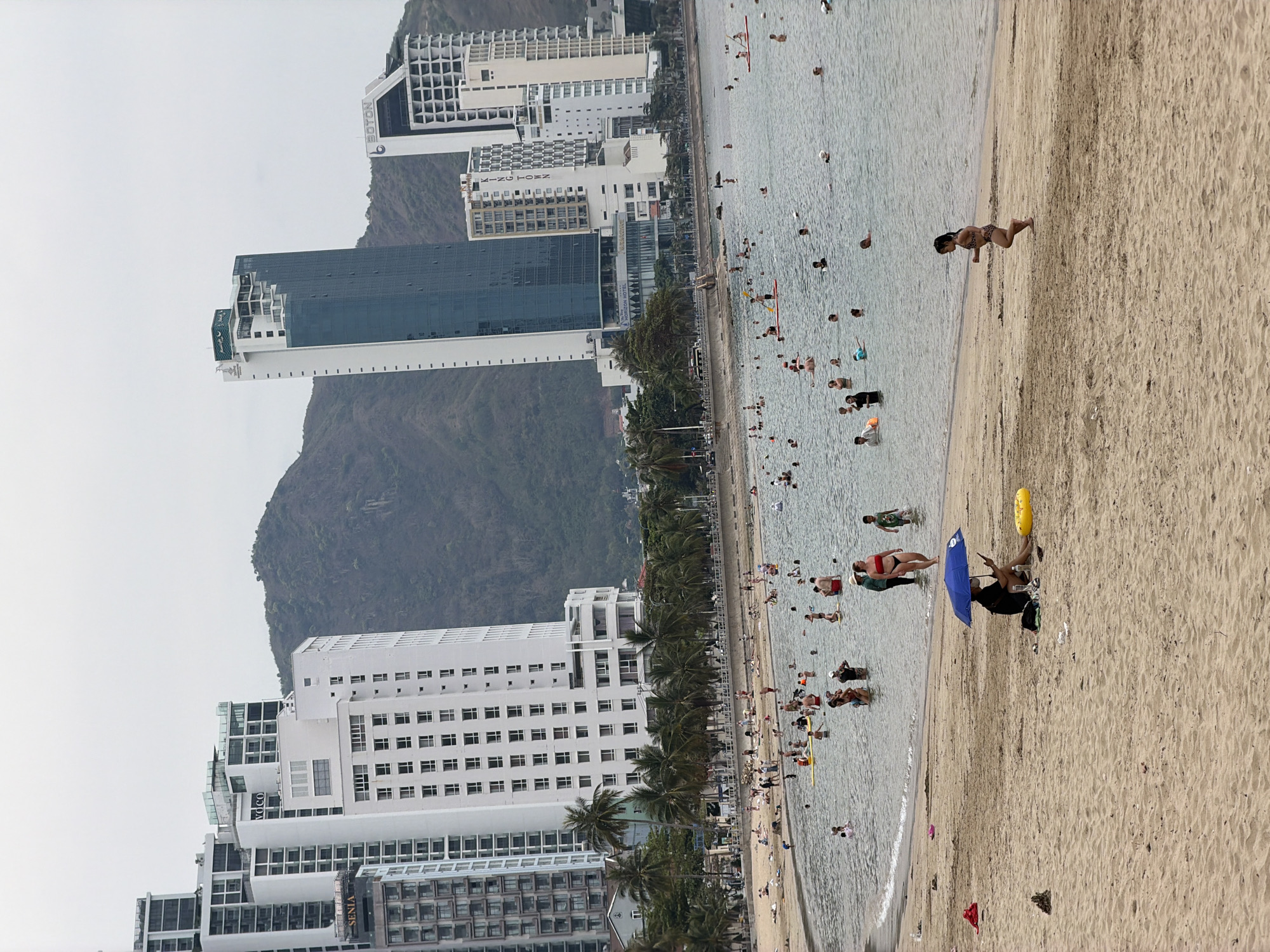 The image depicts a beach scene in Bac Nha Trang Ward, Vietnam. The beach is crowded with people, both in the water and on the sand. Buildings are visible in the background, including hotels and apartments. A mountain provides a backdrop to the cityscape. The sky is overcast. A person sits under a blue umbrella on the beach. People are swimming, wading, and sunbathing. Palm trees line the beach.