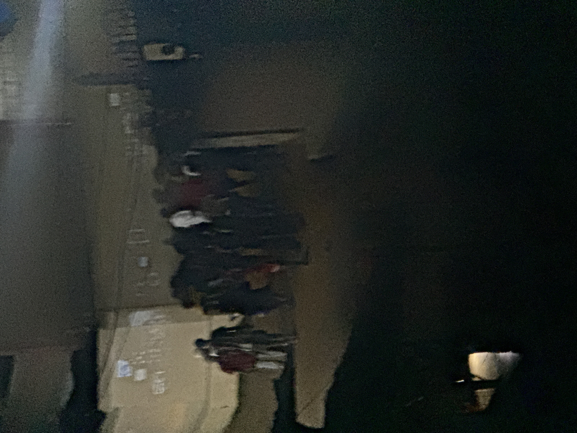 The scene is in Ihiagwa, Nigeria, at night. The image shows a street with a group of people standing on the sidewalk. There are buildings visible on the left side of the street. A streetlight is visible on the right side of the street. The people appear to be engaged in a gathering or conversation. The overall environment is dark, with light sources illuminating portions of the scene. The street surface appears to be concrete or pavement. The building has some signs.
