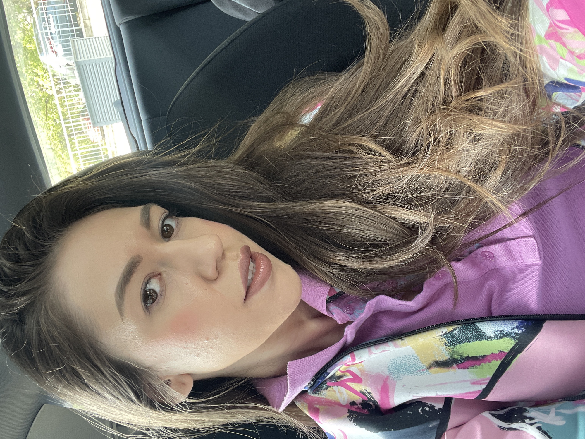 A young woman is depicted in a close-up, slightly tilted portrait, likely a selfie, taken inside a vehicle. She has long, wavy light brown hair with lighter highlights that frames her face and flows over her shoulders. Her dark brown eyes are open, looking towards the camera with a soft, pleasant expression. She has visible blush on her cheeks and a glossy finish on her lips.
She is wearing a pink collared polo shirt, which is layered under an open jacket. The jacket features a vibrant, abstract artistic pattern with prominent colors including pink, green, yellow, black, and white. The interior of the vehicle, visible behind her, appears to be dark-colored.
Through the window on the left side of the frame, a bright outdoor scene is visible. This includes green foliage, a light-colored fence, and part of a building with vertical panels. The bright lighting suggests it is daytime. The image is oriented with the woman's head on the right side, suggesting it was rotated 90 degrees counter-clockwise from a vertical portrait. The location context provided is Sofia, Bulgaria. There is no visible text in the image.