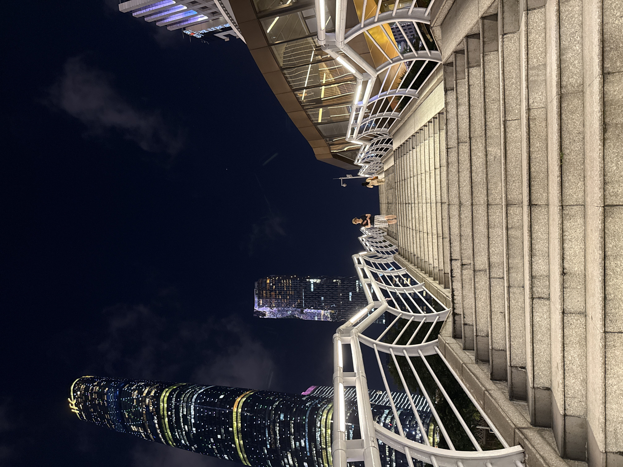 Captured at night in the Tianhe District, China, the image presents a scene featuring staircases. On the steps, two individuals can be observed. The environment is defined by urban elements, including buildings and a dark sky with clouds. The overall lighting enhances the details of the architectural structures and objects present in the frame.
