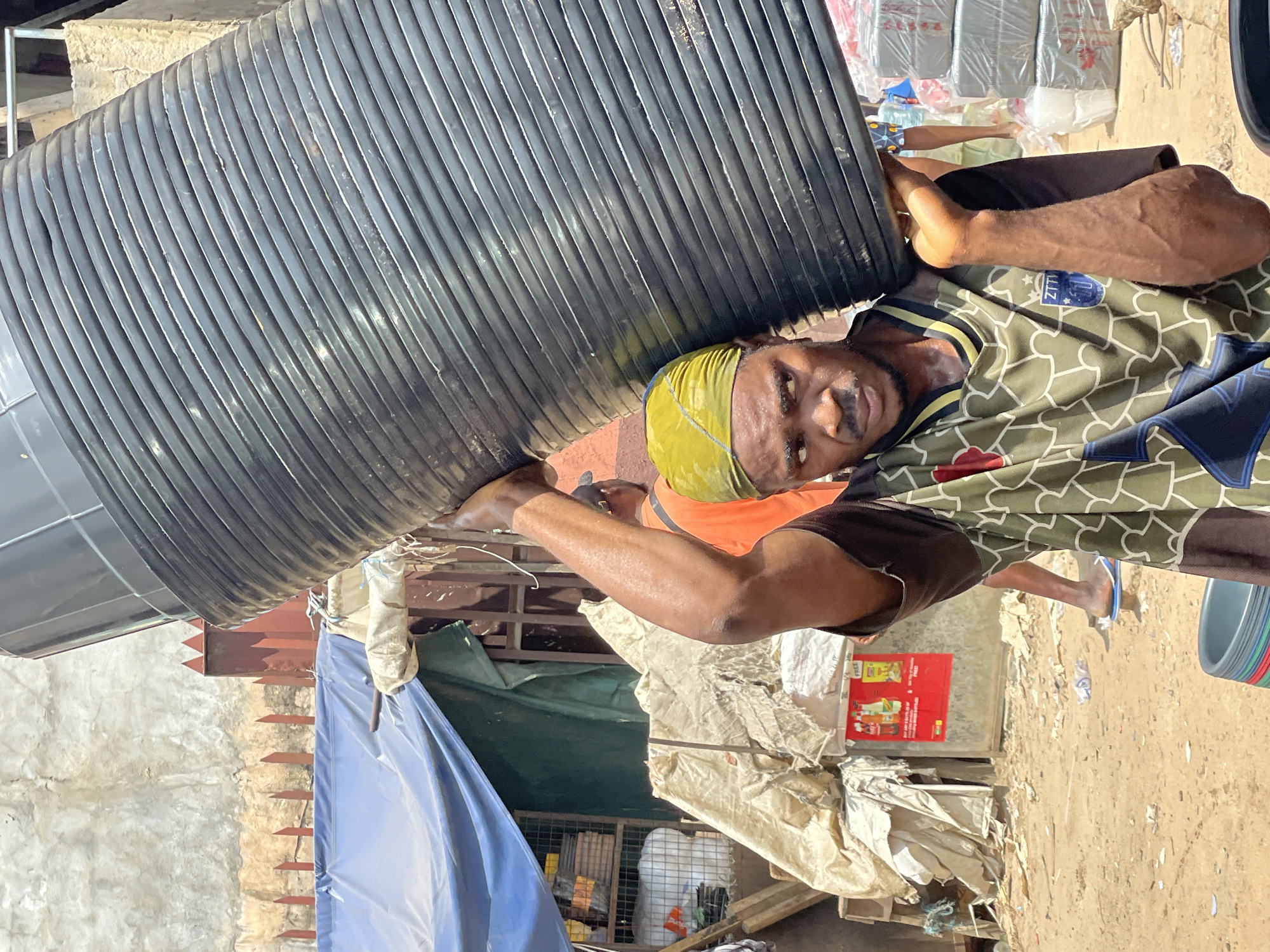 A man with dark skin and a yellow headwrap is carrying a large, black, ribbed cylindrical object horizontally across his shoulders. He is wearing a green and brown patterned t-shirt with a blue emblem on the left sleeve. The man is looking to the left with a serious expression. In the background, there are stacks of white plastic-wrapped items, a red sign with text and images, and blue and green tarps draped over structures. The ground appears to be dirt and there are some scattered debris and items. The scene suggests an outdoor market or storage area.