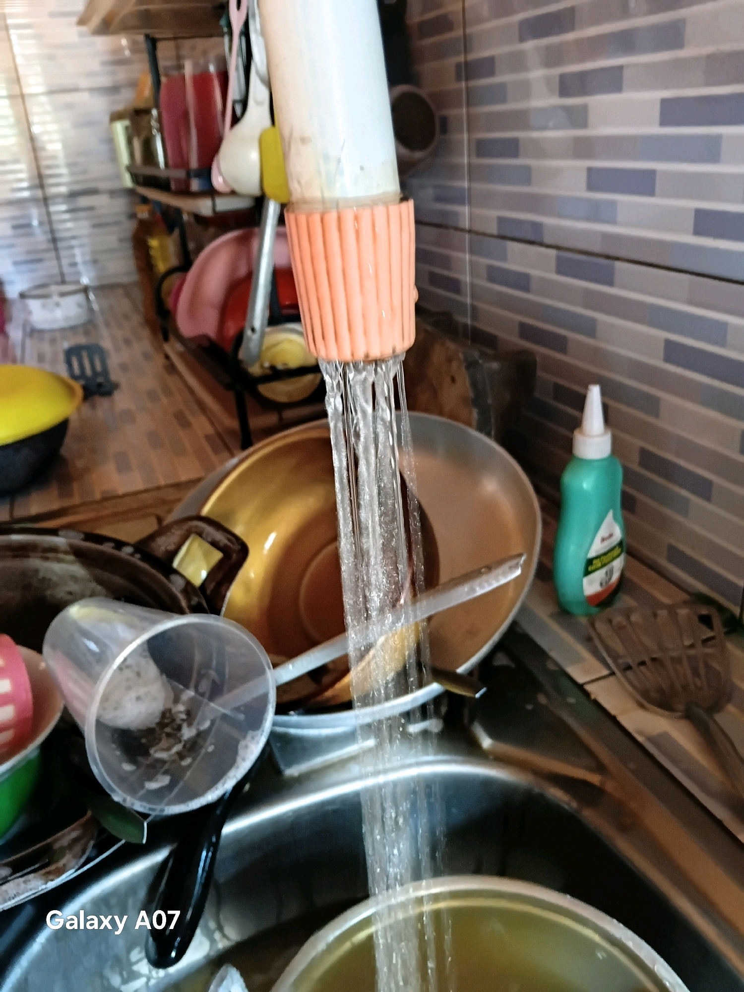 The image captures a close-up, eye-level view of a kitchen sink in Margo Rukun, Indonesia. Water is actively running from a faucet, which has an orange, ridged attachment, into a sink filled with various dishes and kitchenware. In the foreground, a clear plastic cup with some debris and foam is visible, partially submerged in the sink. To the right of the faucet, a green bottle with a white cap sits on the countertop, and behind it, a tiled backsplash with a pattern of grey and blue rectangles is present. The overall impression is of a busy, functional kitchen environment with dishes being washed. The lighting suggests an indoor setting, likely during the day, with light coming from an unseen source, possibly a window or overhead lights.