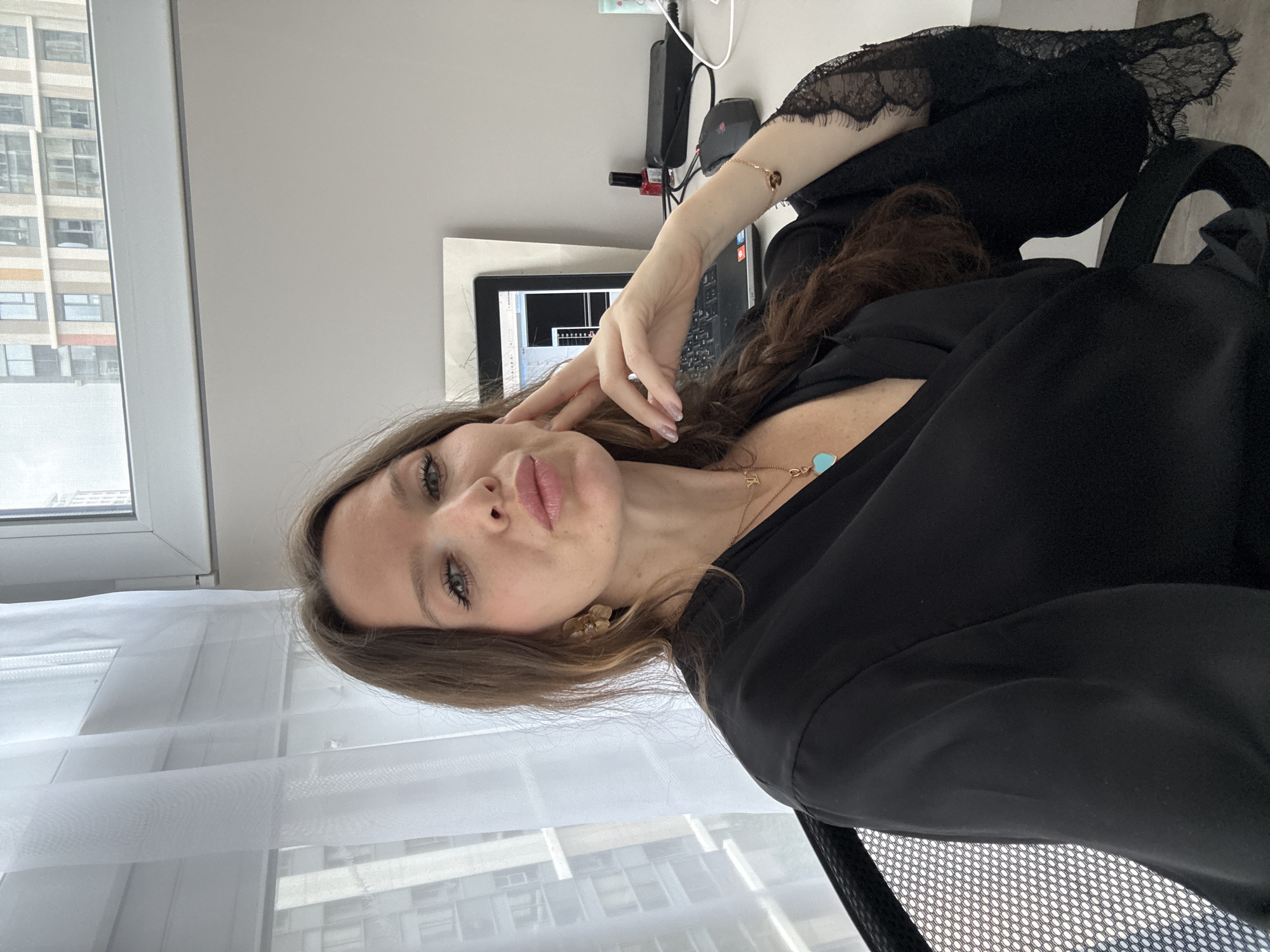A woman is in a room. She is seated facing the camera. Behind her is a window. Outside the window is a building. According to the windows and building the location is likely in Moscow, Russia. The woman is wearing a black top with lace. She is wearing gold jewelry. There is a laptop and electronics on the desk.